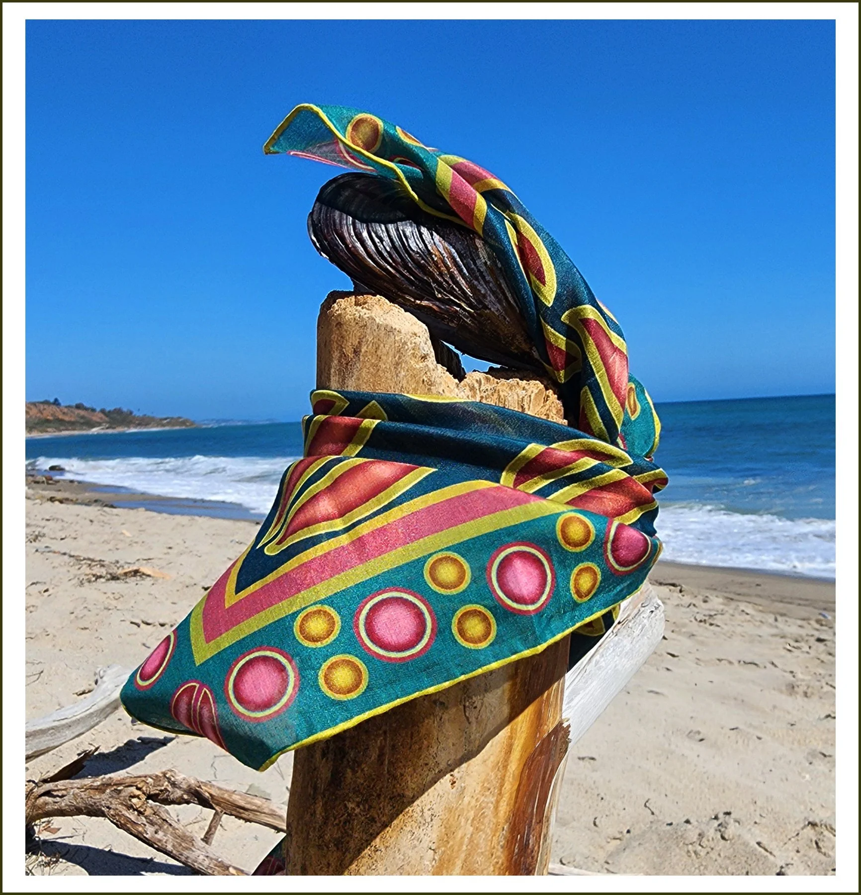 A wooden sculpture with a colorful scarf wrapped around it, located on a sandy beach with the ocean and clear blue sky in the background.