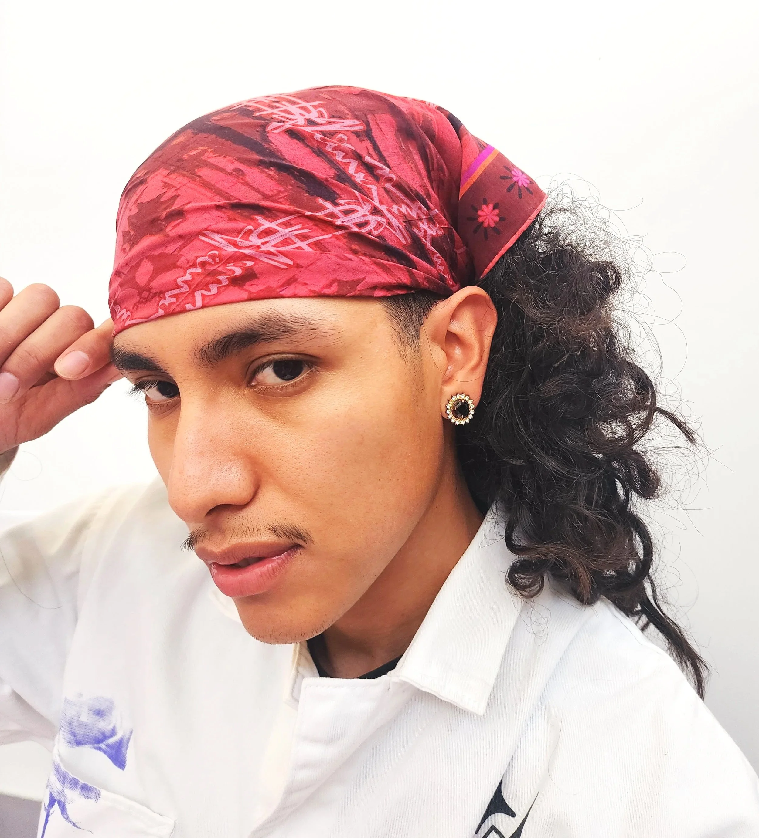 Person wearing a red bandana, white shirt with logo, and a black earring, posing with one hand saluting on a white background.