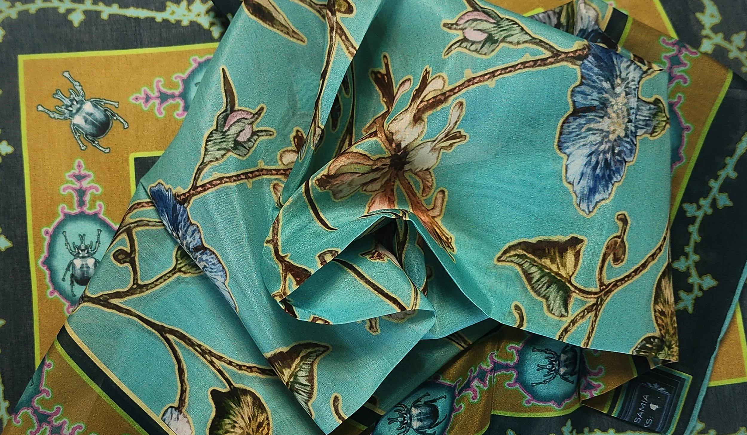 Close-up of a turquoise silk scarf with floral and insect patterns, draped over a yellow and black printed fabric background.