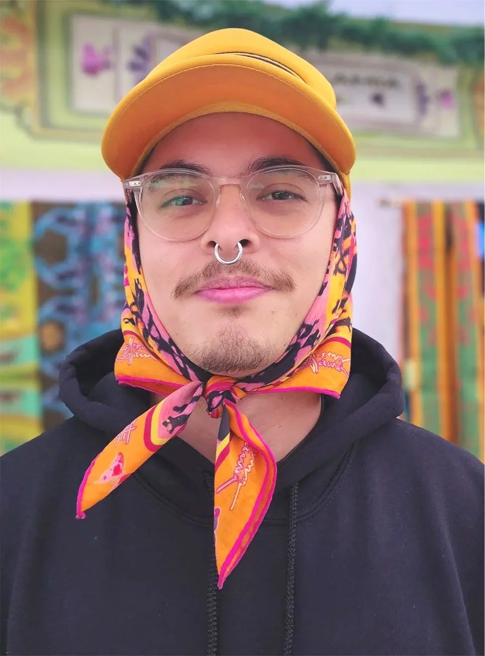 A young man with glasses, a septum piercing, and a colorful scarf tied around his head. He is wearing a yellow cap and a black hoodie, standing in front of colorful fabric or artwork.