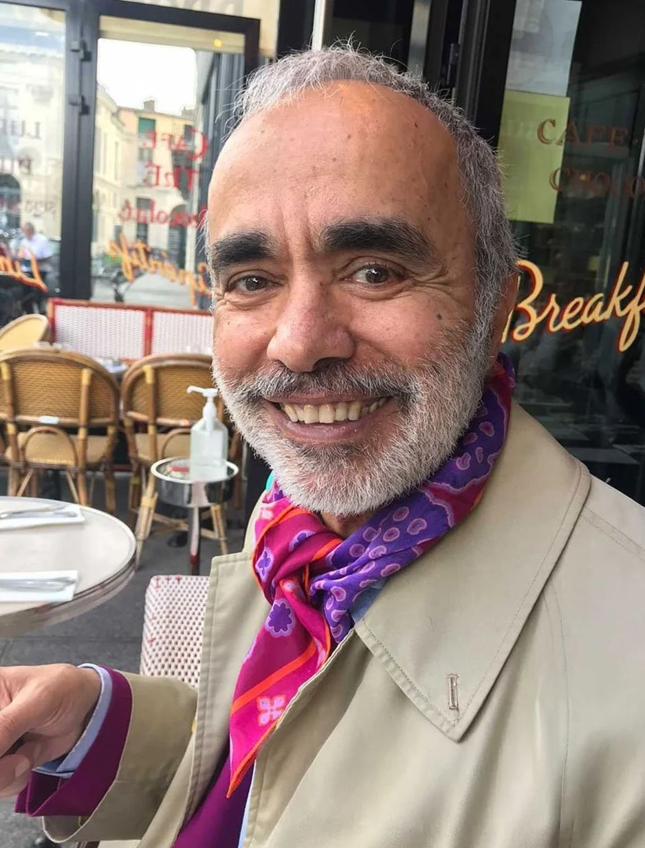A smiling man with gray hair and beard, wearing a beige coat and a purple and pink patterned scarf, sitting at an outdoor cafe.