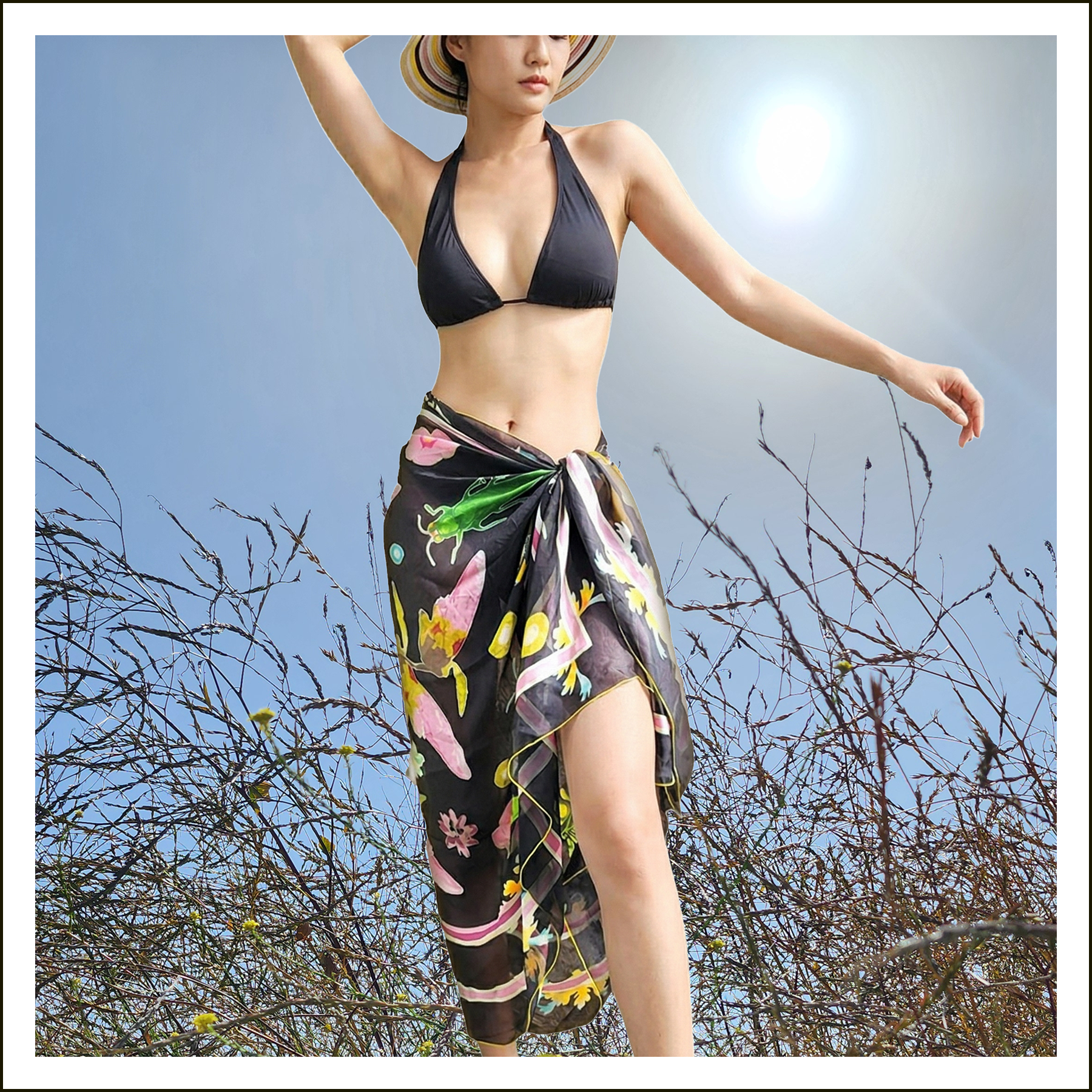 A woman wearing a black bikini top, a wide-brimmed hat, and a floral sarong standing among dry, leafless branches with a bright sun and blue sky in the background.