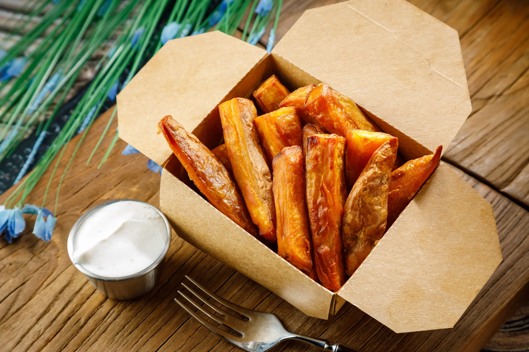 Crispy fried eggplant sticks in a brown paper box on a wooden board, served with a side of white dipping sauce in a small plastic cup.