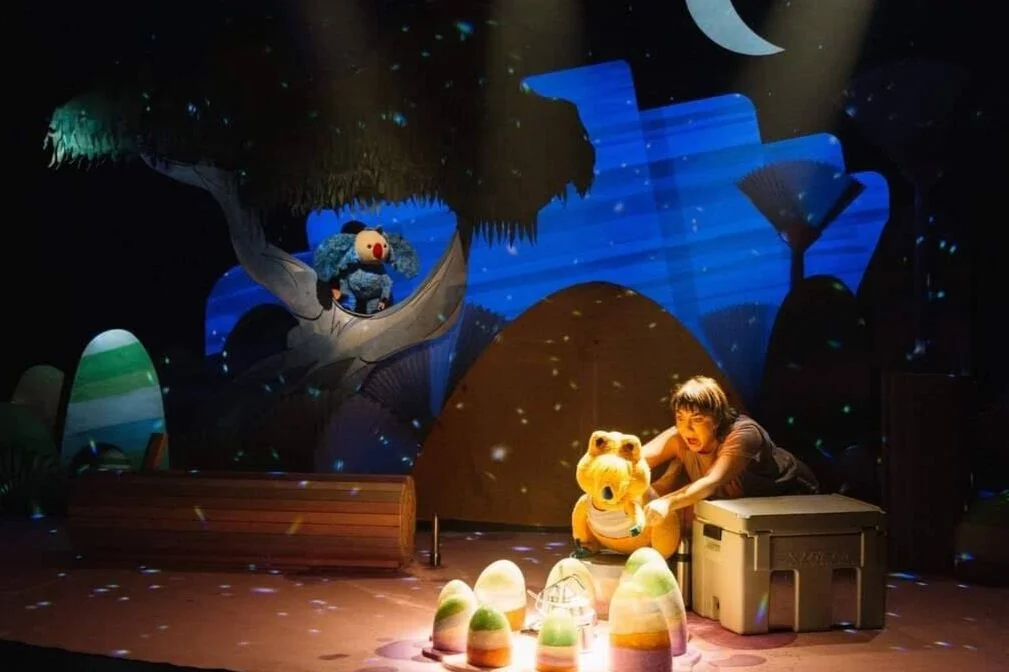 A theatre stage with pastel coloured set pieces making up a stylised portrayal of the Australian bush. There is a sharp spotlight over the campsite, and a yellow Quokka puppet leaning into it. Chiara is behind the puppet with an excited face.