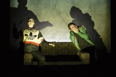 Chiara dressed as Maz in an adidas jacket sits on the left side of a couch, while Jim Smith as Lee sits on the right.  Theire faces a lit from the bottom,.