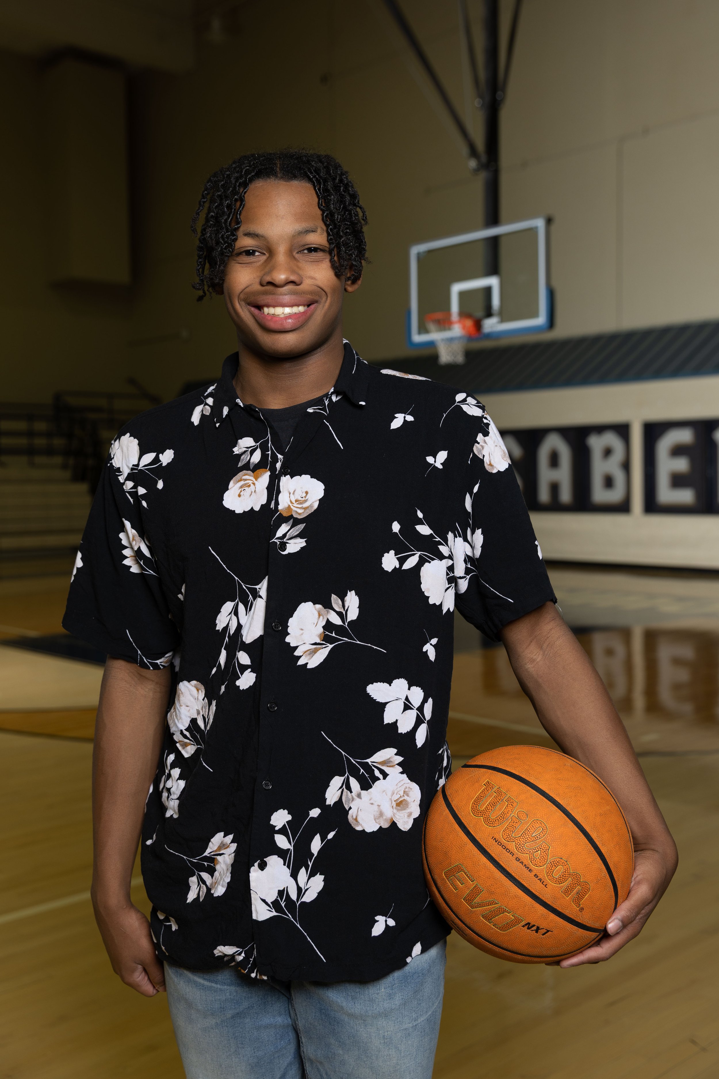 A young man with curly hair wearing a black floral shirt and light blue jeans, standing in a gymnasium with a basketball in his right hand.