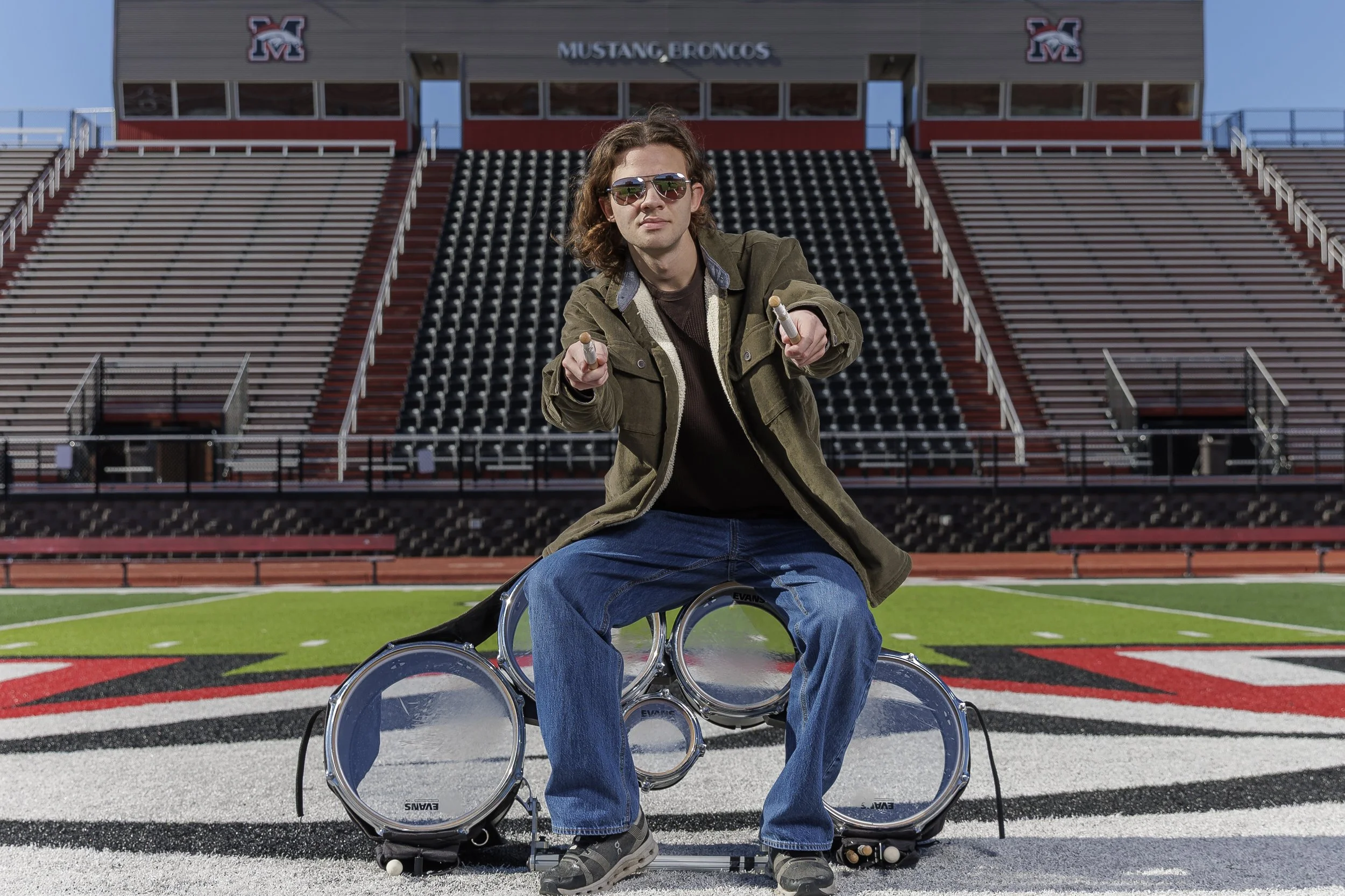 A young man with long curly hair wearing sunglasses, a brown shirt, and a green jacket  He is pointing with both hands towards the camera. Behind him is empty bleachers and a stadium with a sign that reads 'Mustang Broncos' and the initials 'M' in red, black, and white on the building.