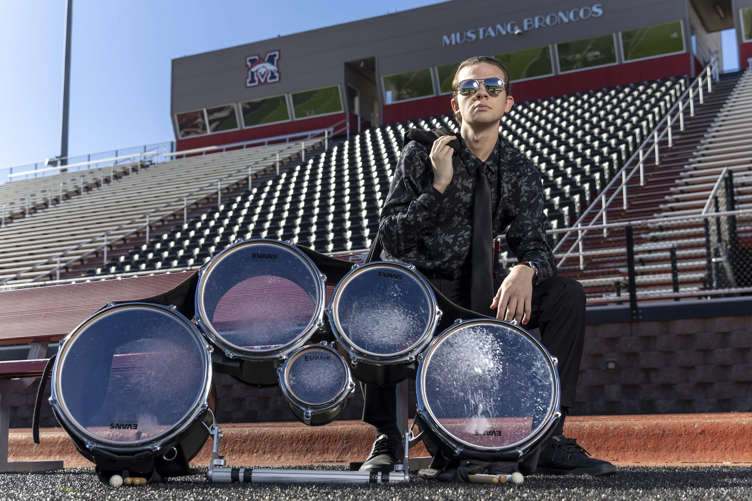 A high school senior male student in sunglasses sitting on a football stadium seat with a set of percussion drums in front of him. The stadium has empty seats and signage that reads 'Mustang Broncos'.