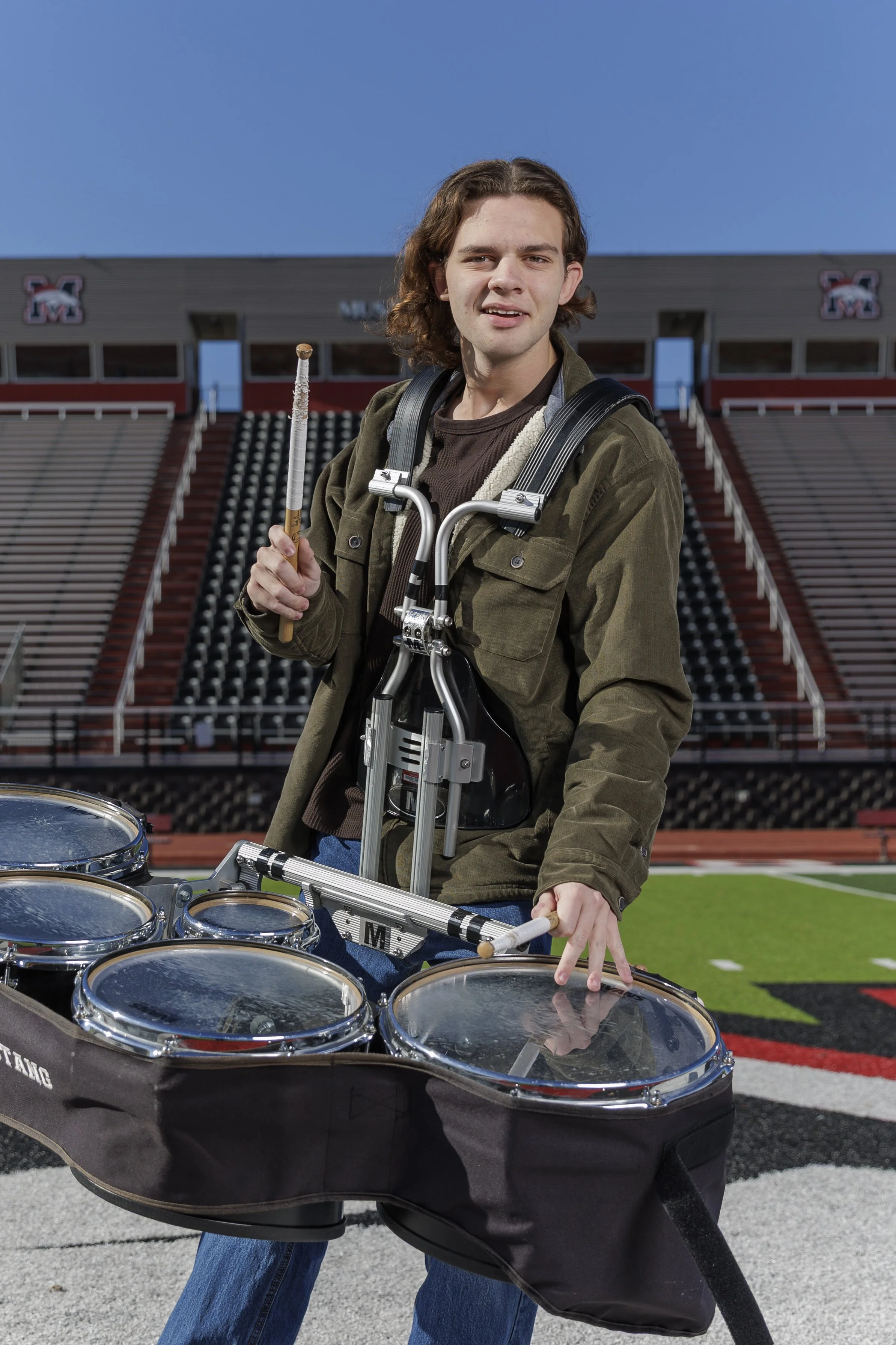 Young man with long brown hair standing with a drum set in a stadium, holding drumsticks, wearing a green jacket, and standing on a football field.