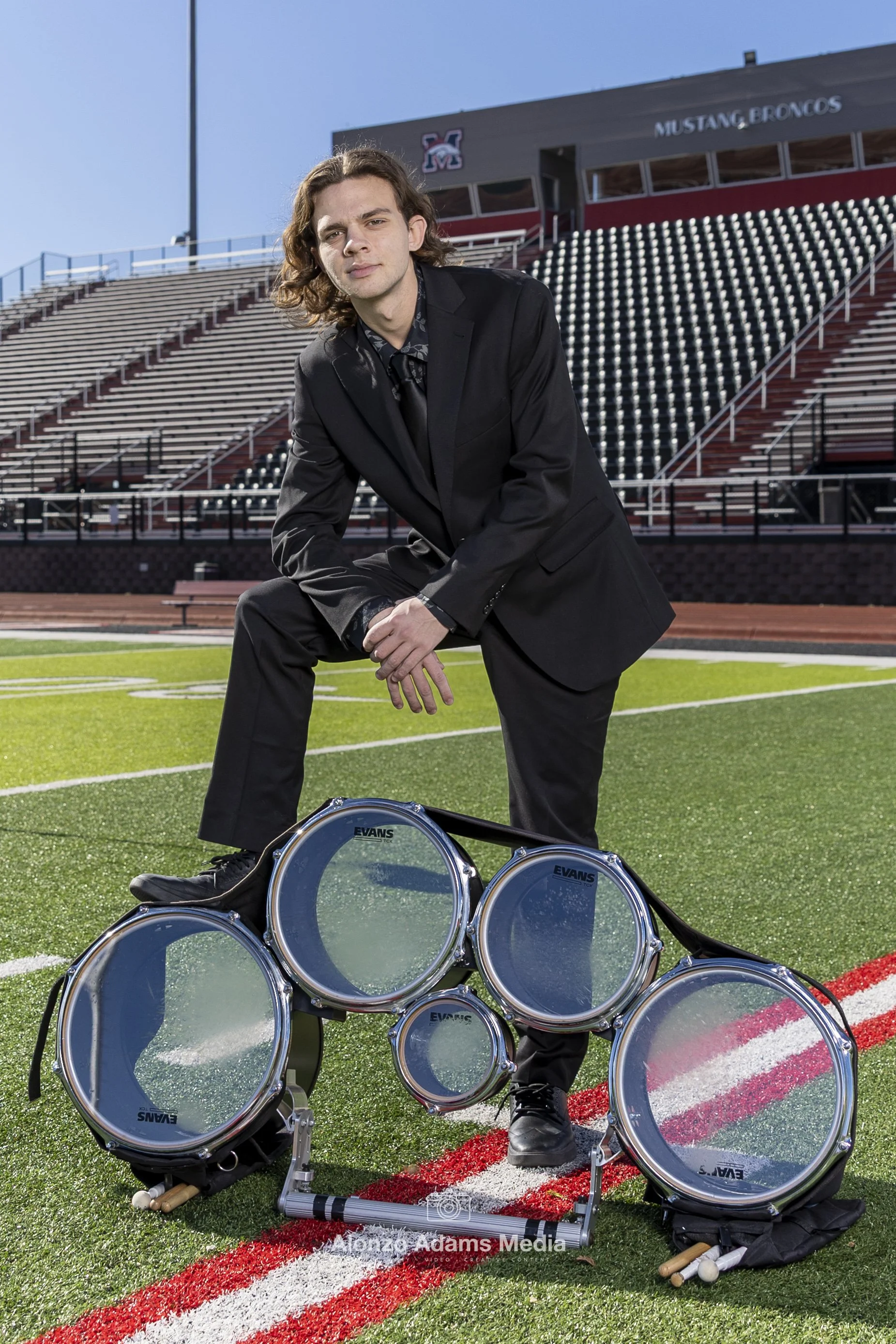 Man in a black suit kneeling on a football field, surrounded by drum sets, in front of bleachers and stadium stands, with a building in the background displaying 'Mustang Broncos'.
