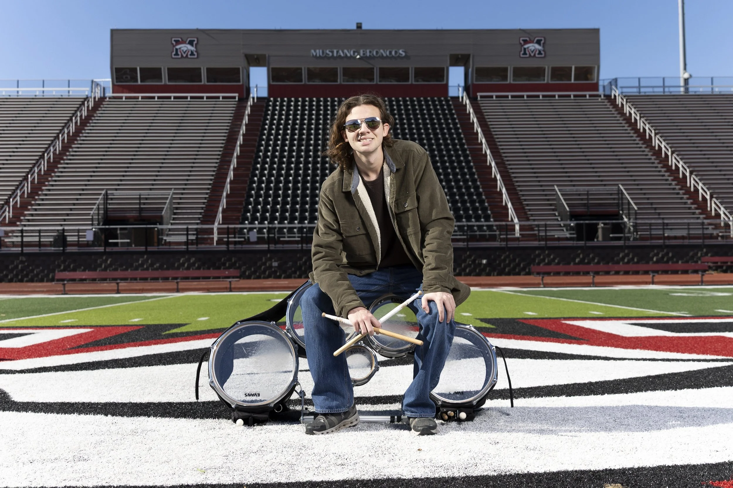 A young high school senior in sunglasses sitting on a drum set in the middle of a football field with bleachers in the background, at a stadium named Mustang Broncos.