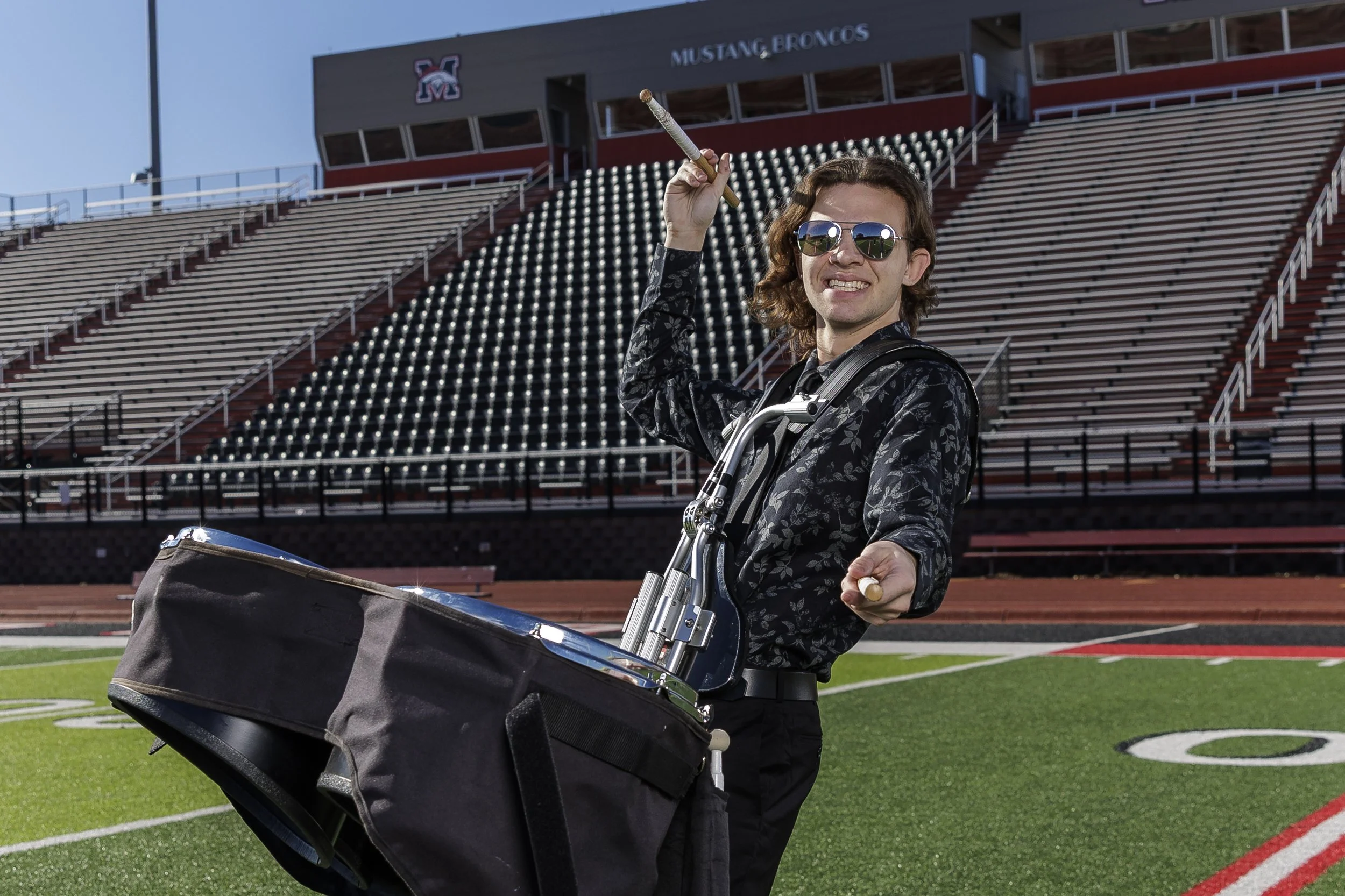 A man with curly hair wearing sunglasses and a patterned shirt is standing on a football field, holding a drumstick in one hand and smiling. He is positioned behind a large percussion drum with a bag attached, in front of empty stadium bleachers. The stadium has a sign that reads 'Mustang Broncos' and features a logo with the letters 'SM'.