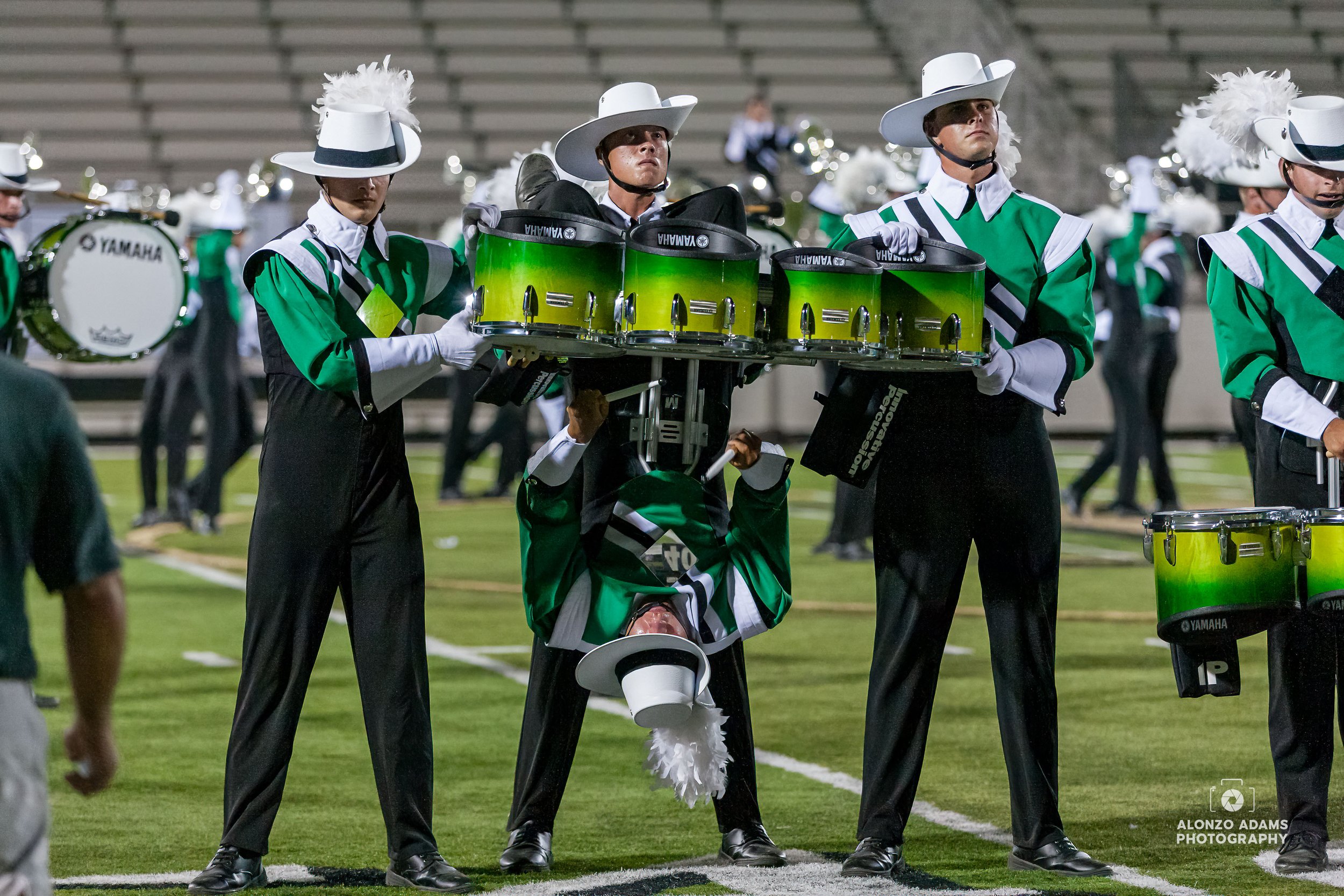 two drum corps members hold a percussion drummer upside down with his drum on while he plays during a performance.