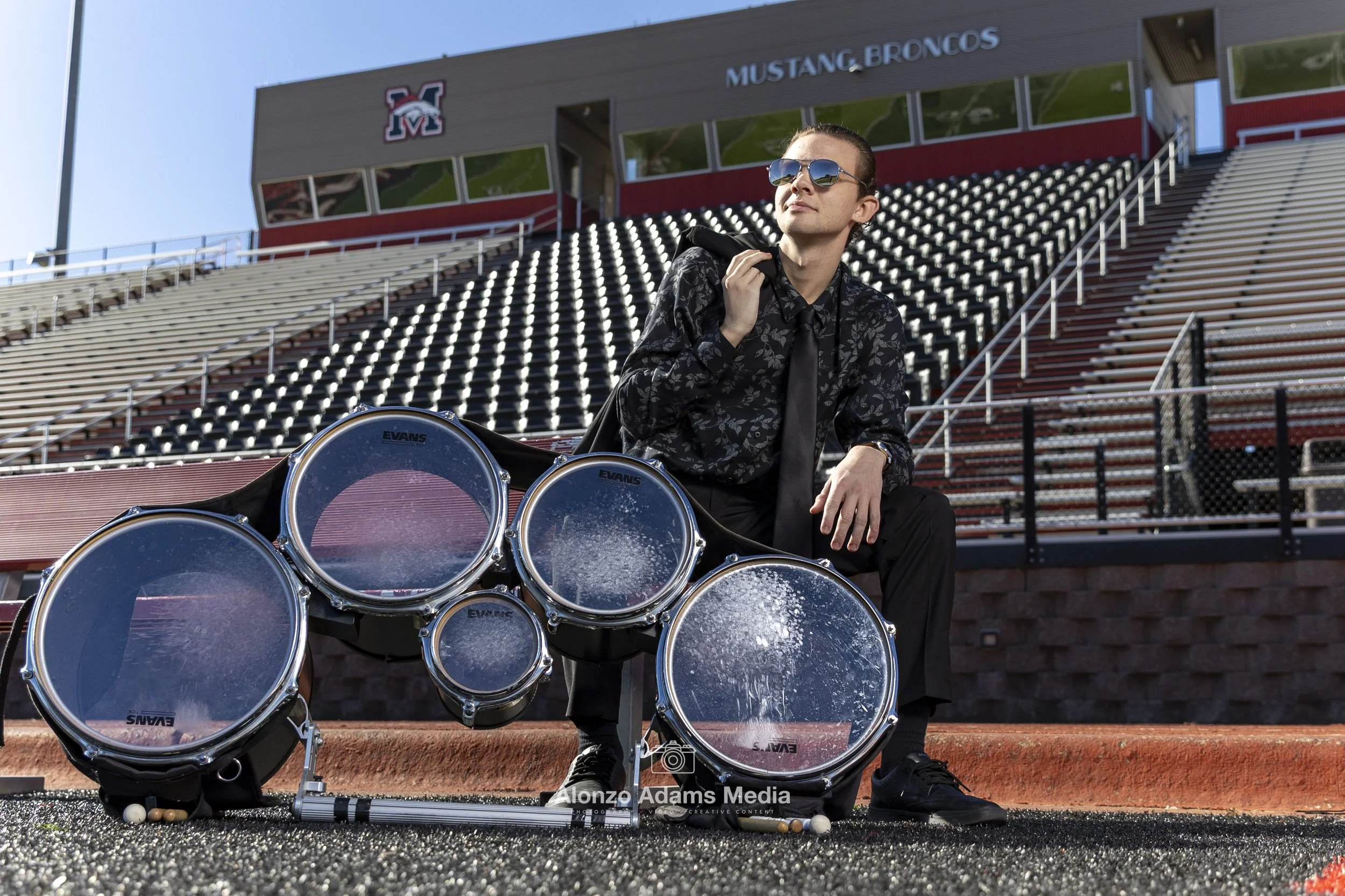 A man in sunglasses and dark clothing sits on the steps at a football stadium, with a set of marching band drums in front of him.
