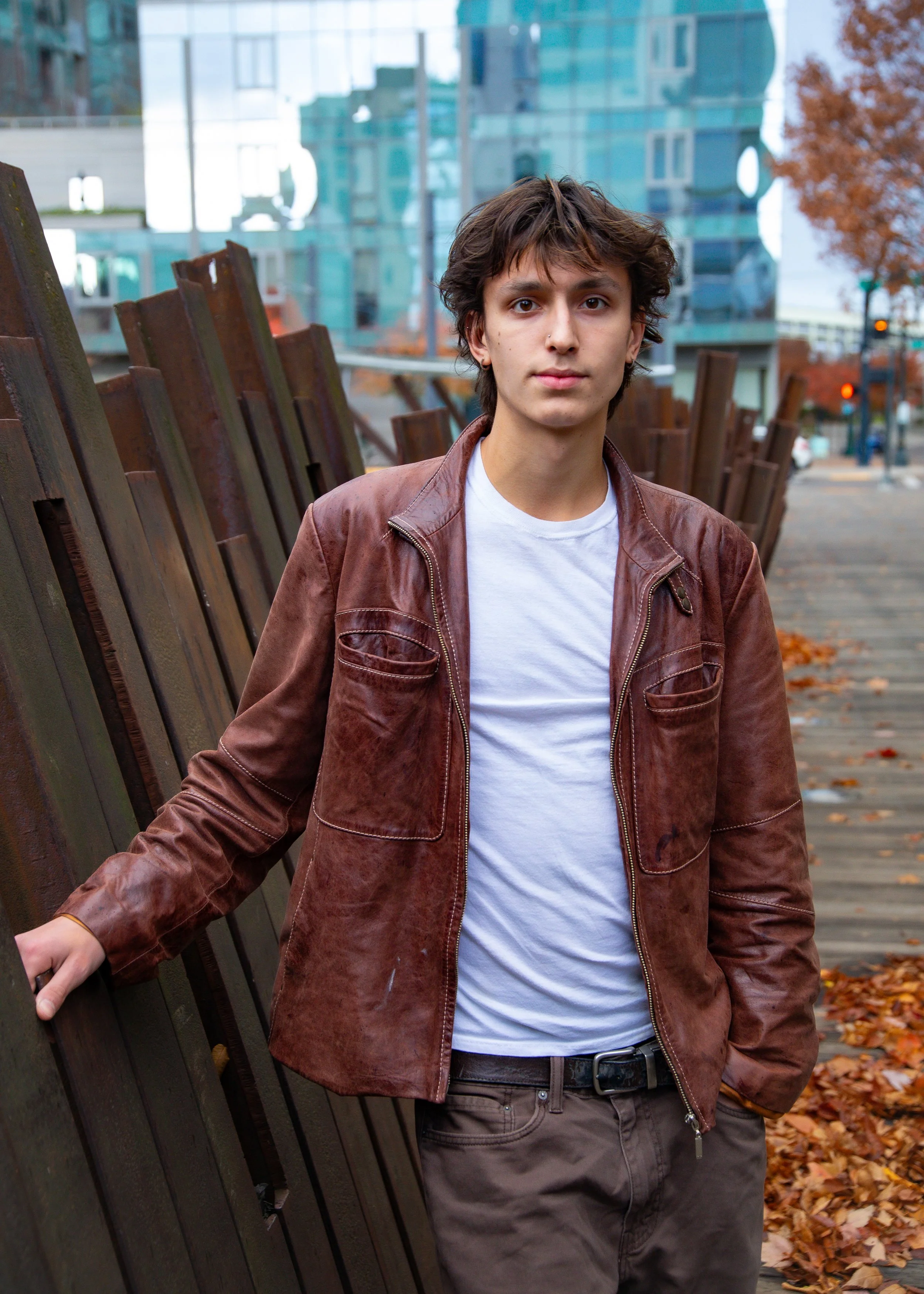 Young man wearing a brown leather jacket and white t-shirt standing outdoors on a wooden path with autumn leaves and modern buildings in the background.