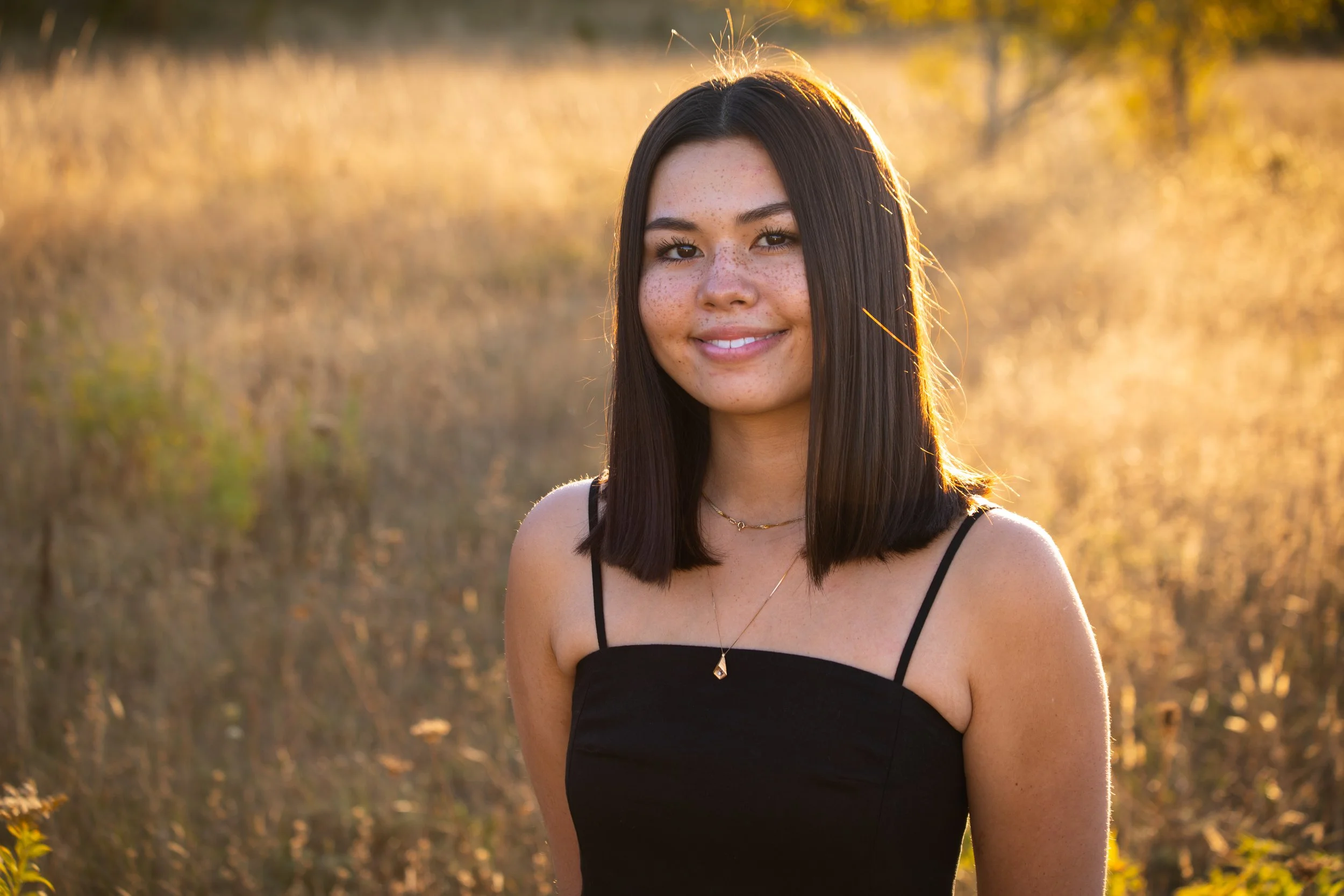 Young woman with shoulder-length hair smiling in a sunlit field, wearing a black top and necklace.