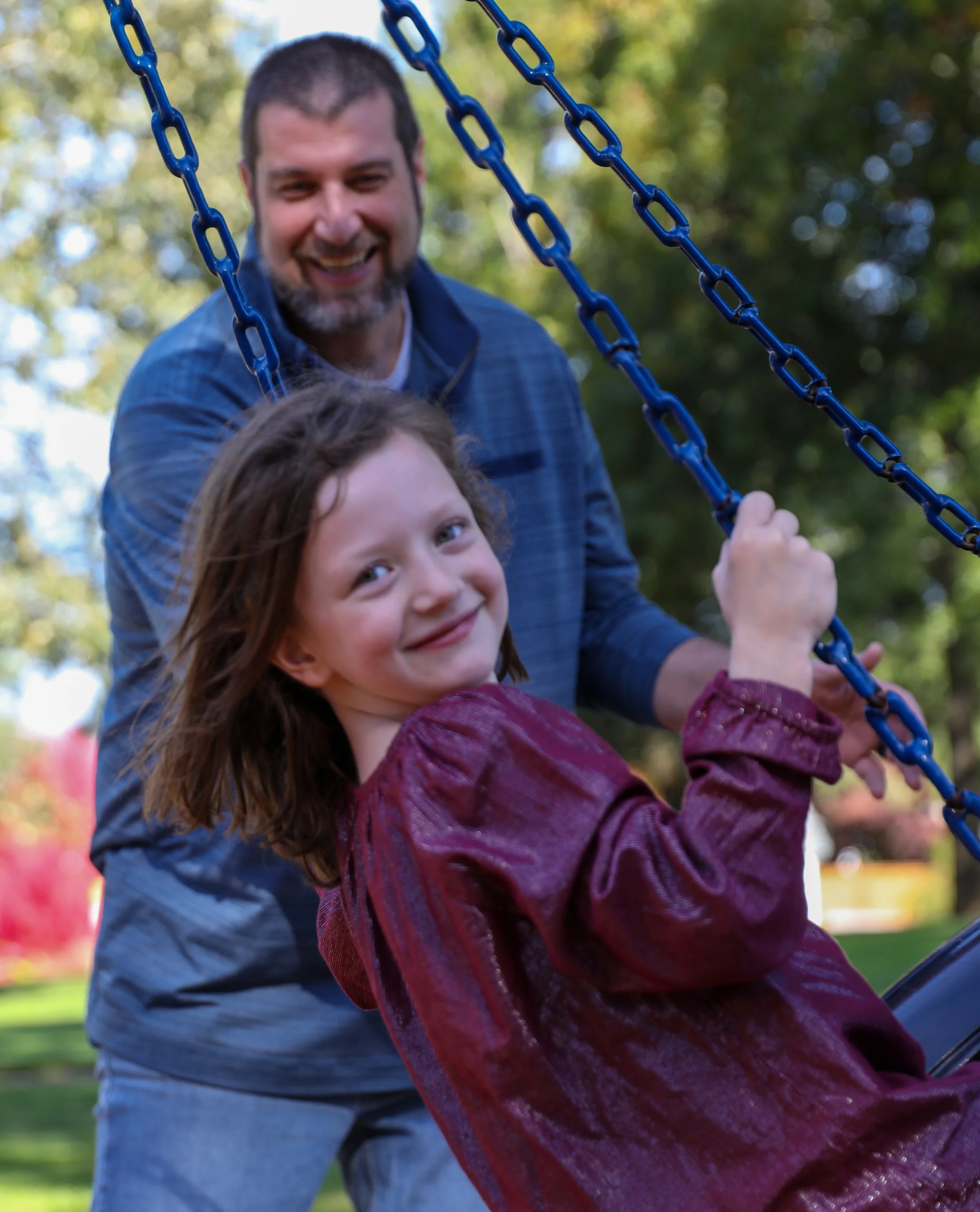 A smiling child on a swing being pushed by an adult in a park, surrounded by greenery.