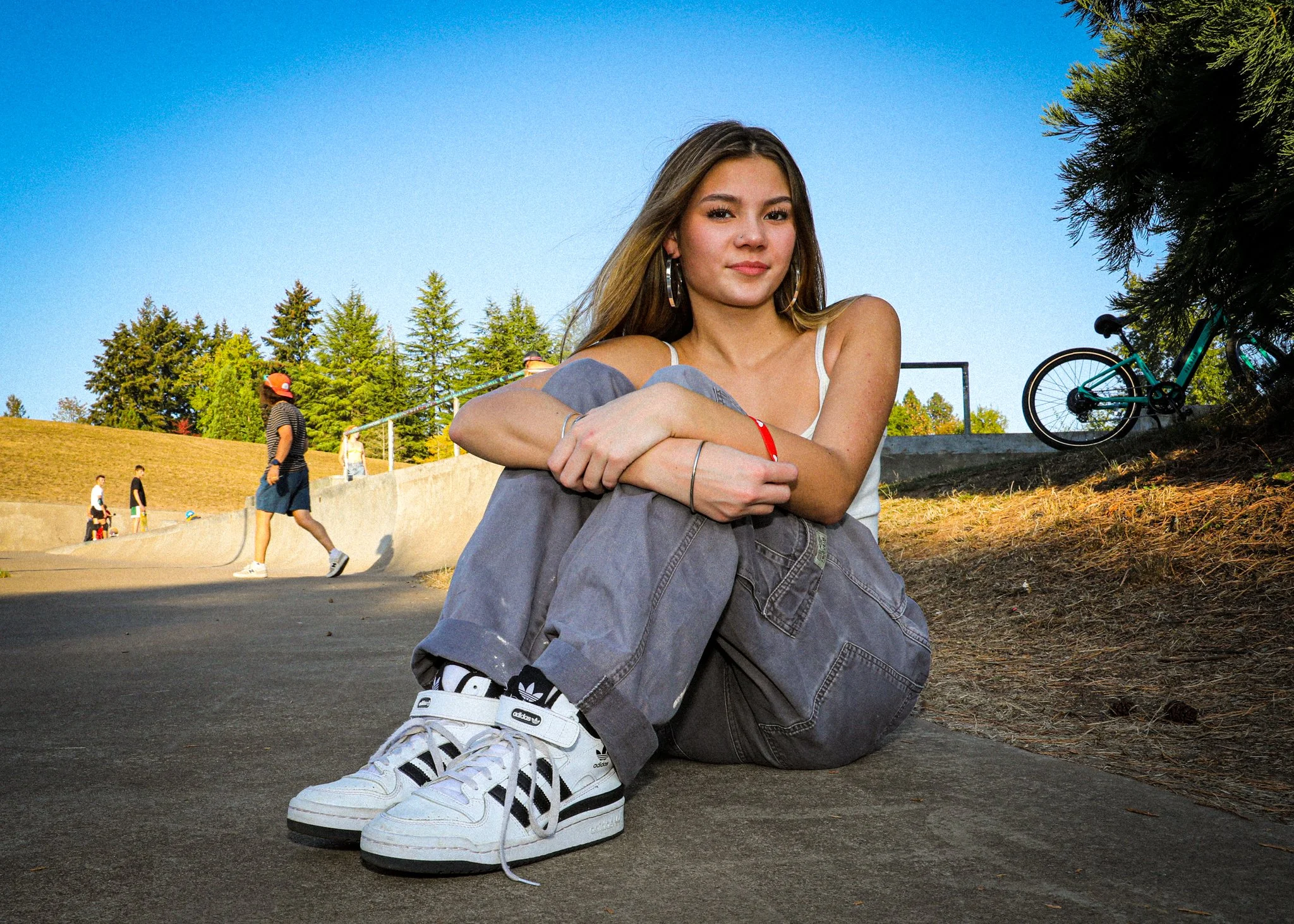 Young woman sitting on the ground at a skate park, wearing jeans and sneakers, with people walking and a bicycle in the background.