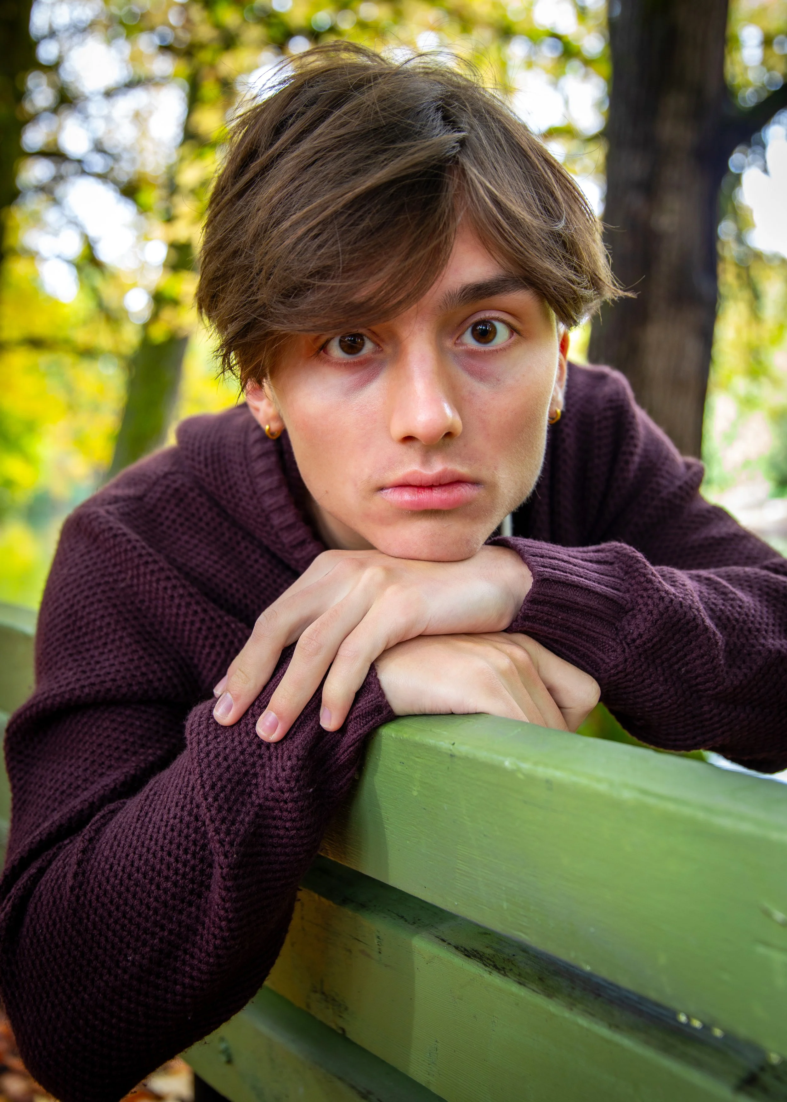 Young person with brown hair leaning on a green bench outdoors, wearing a maroon sweater, with a blurred natural background.