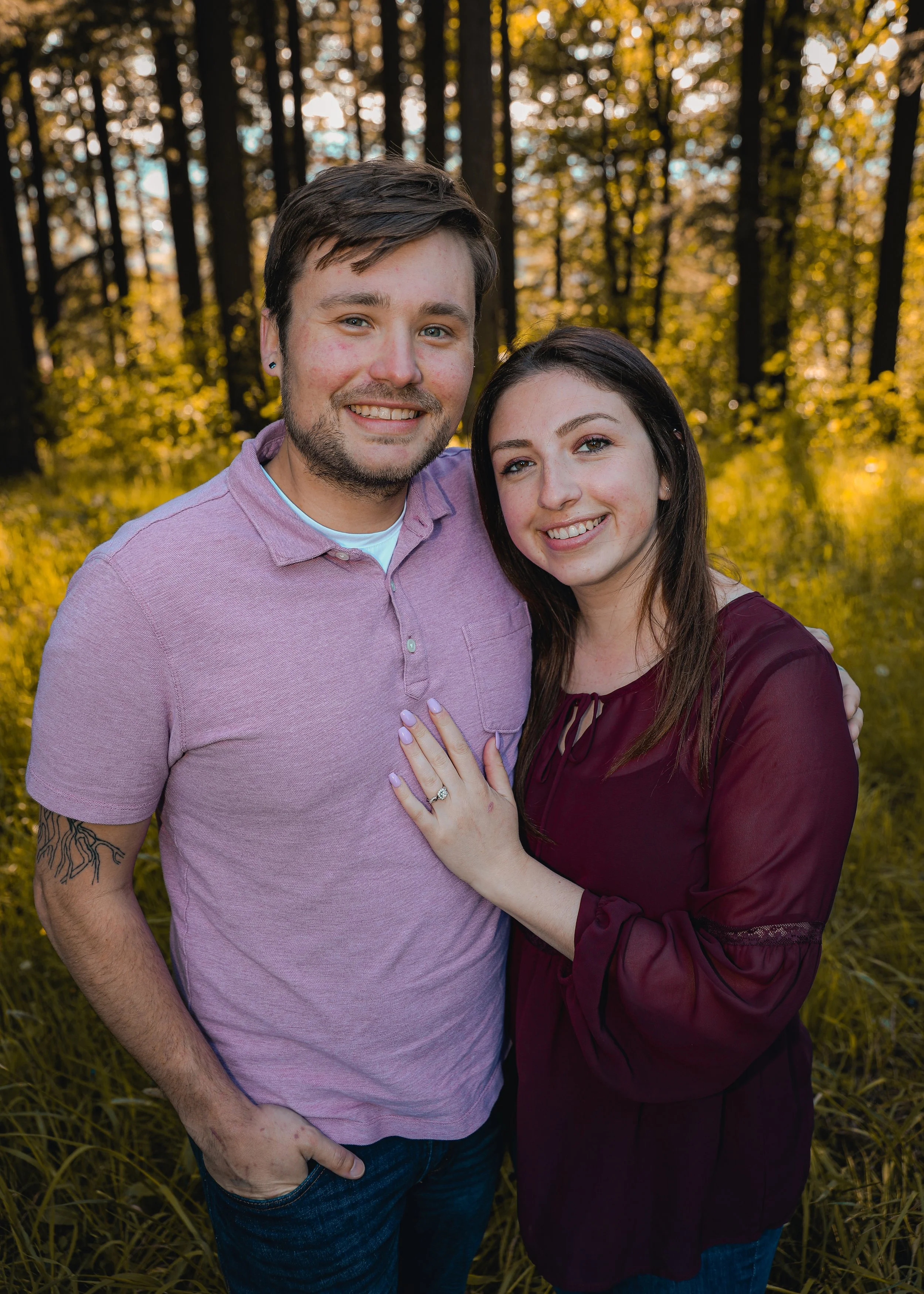 A couple smiling and posing in a forest with the woman showing her engagement ring.