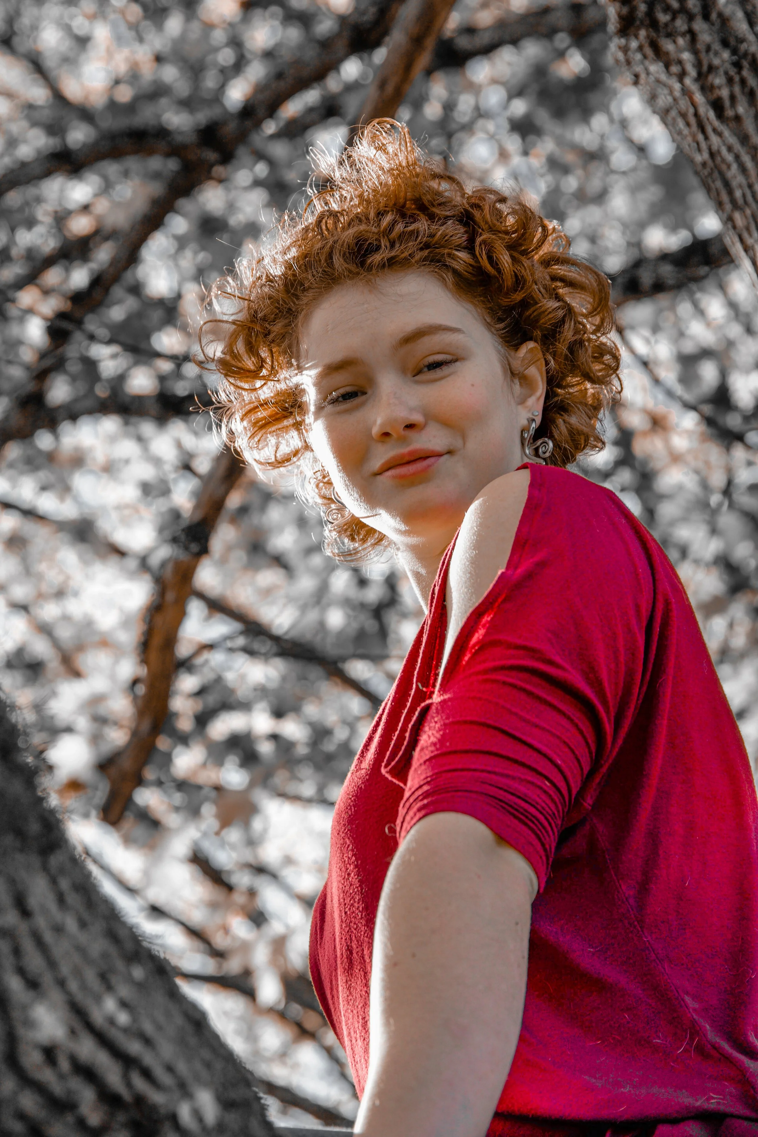 A person with curly hair wearing a red shirt, standing outdoors by trees.