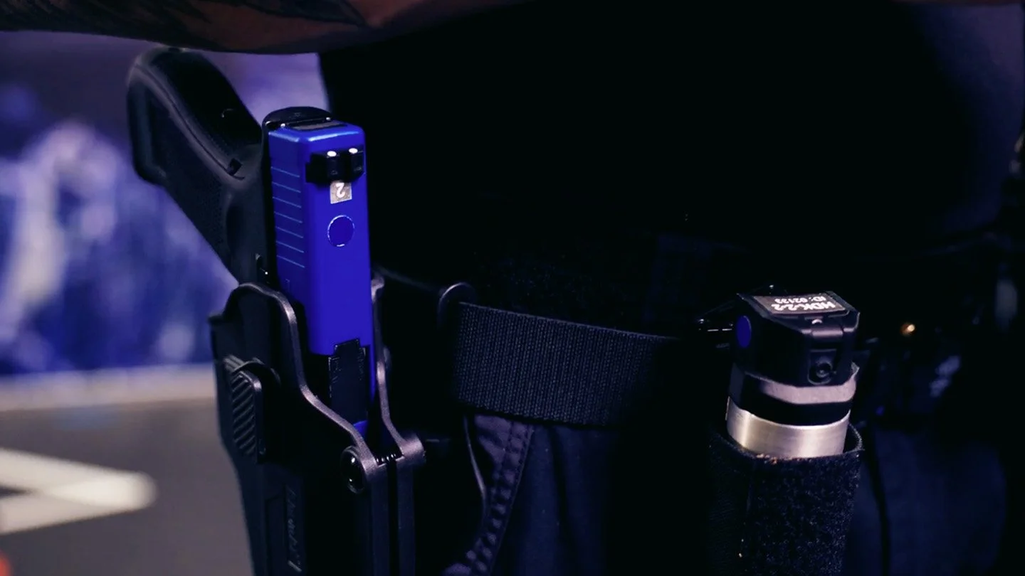 Close-up of a police utility belt featuring a blue training gun in a holster and a small canister next to it.