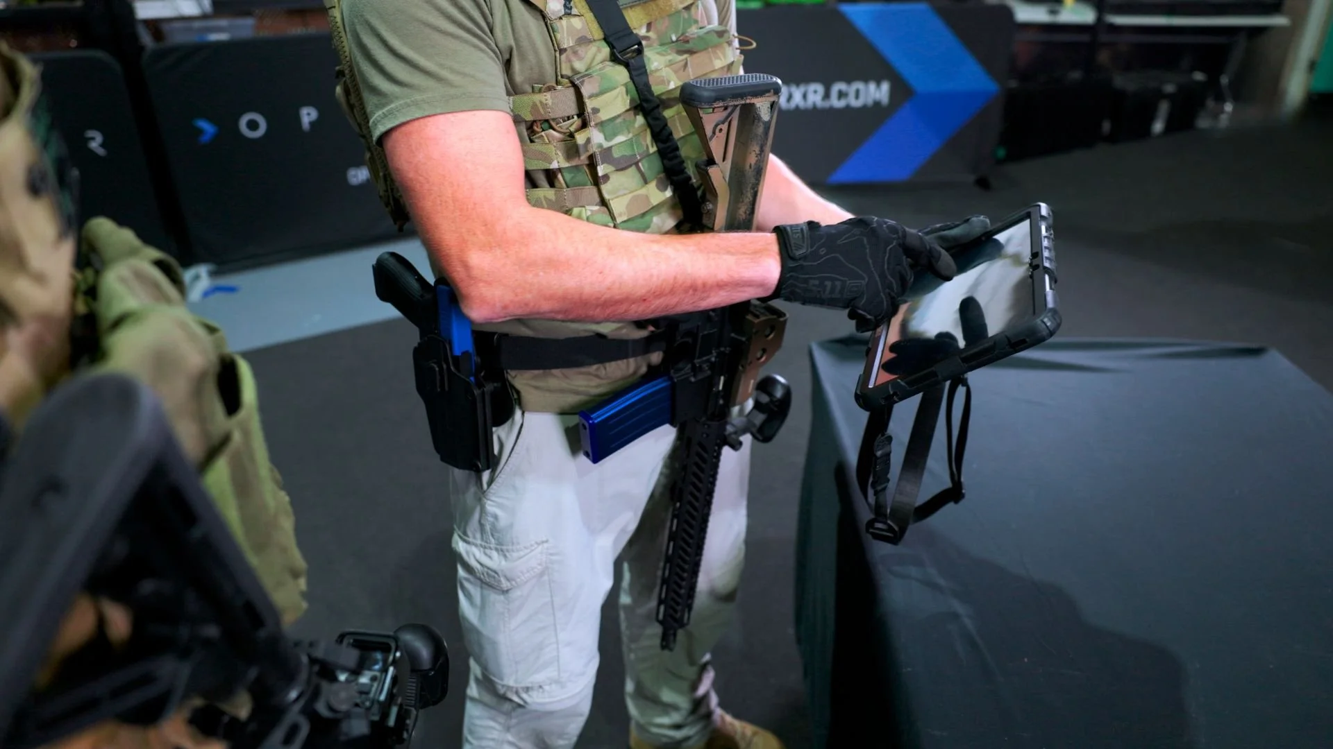 A person dressed in tactical gear, wearing a camouflage vest and black gloves, is holding a tablet device, standing near a table with a black cover at an indoor event or exhibition.