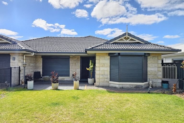 Front view of a house with a stone exterior, security roller shutters, and a tiled roof. There are potted plants and a well-maintained lawn in the front yard under a partly cloudy sky.