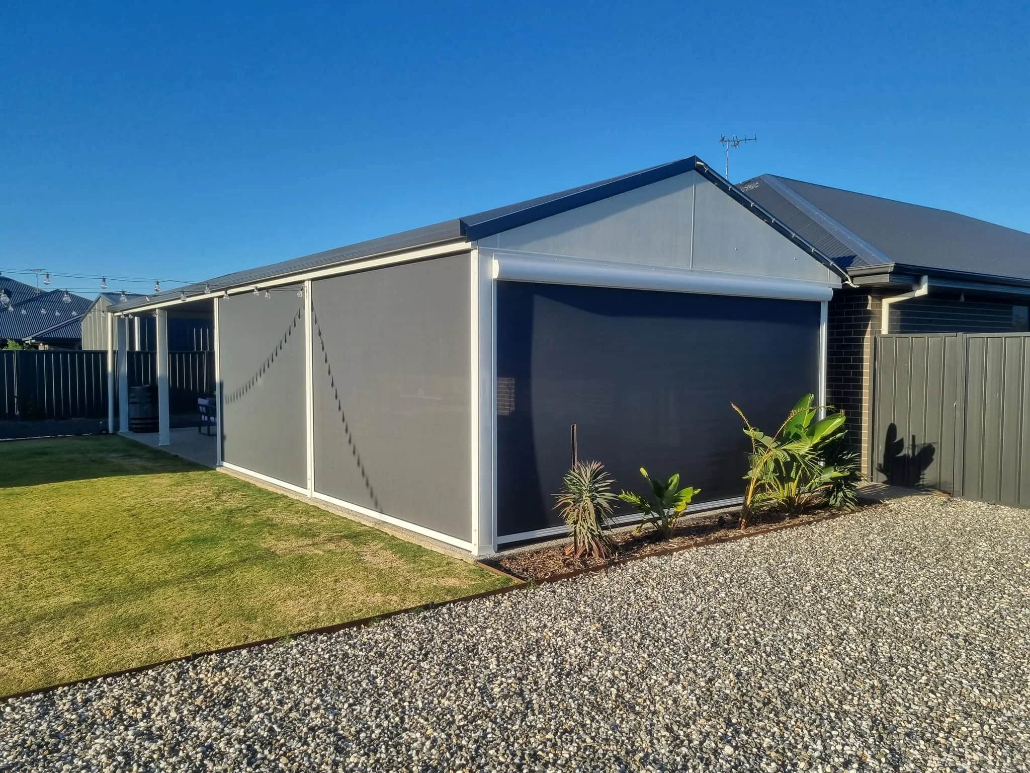 Backyard with a small lawn, gravel path, and a screened-in carport using focus outdoor blinds attached to a house with a grey roof and brick wall.