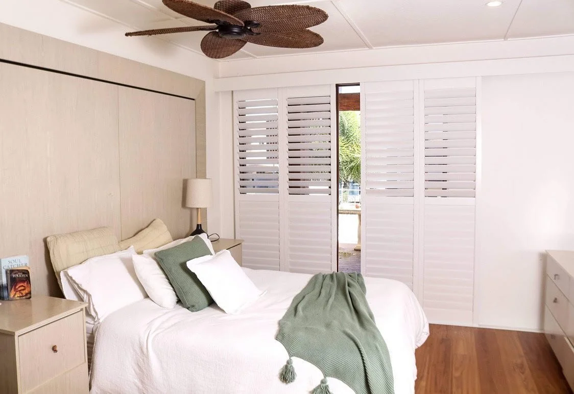 A bedroom with a bed, pillows, a bedside table with a book, a lamp, a ceiling fan, and white plantation shutters on the window.