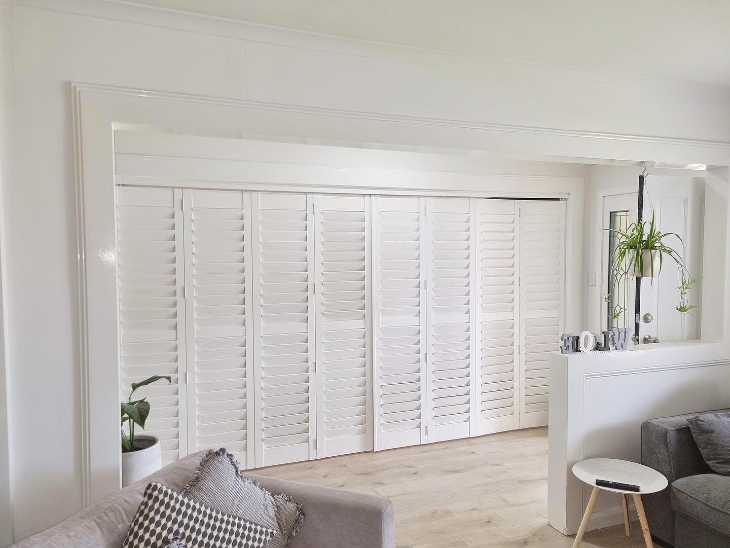 Living room with white plantation shutters, a grey lounge with patterned cushions, a small white side table with a remote, a potted plant, and a decorative 'WILD' sign on a ledge, with a hanging plant nearby.