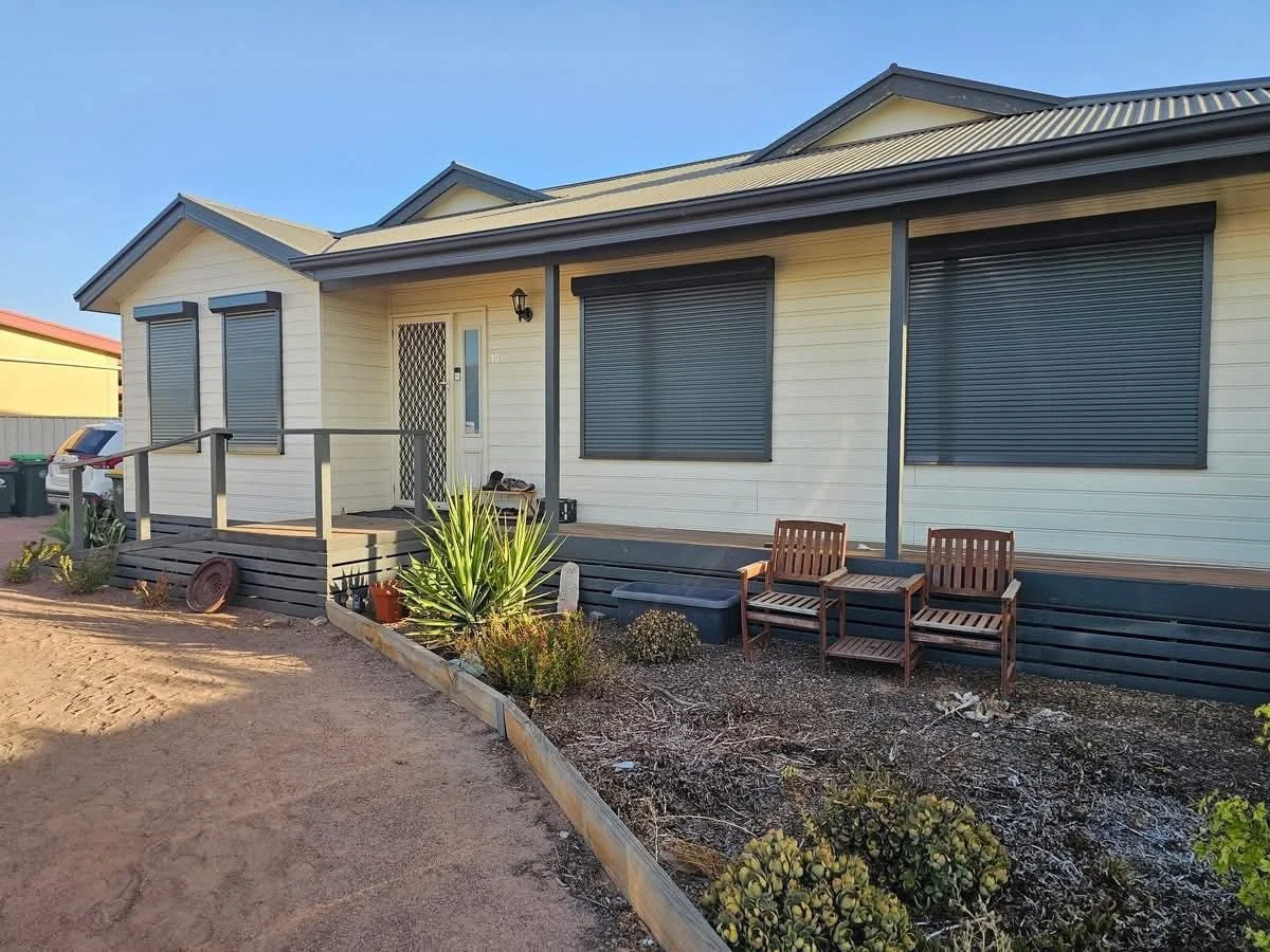 A Port Augusta house with closed black security roller shutters on the windows and a small front porch with a railing. There are two wooden chairs and a small table in the garden area, which has plants and a dirt pathway.