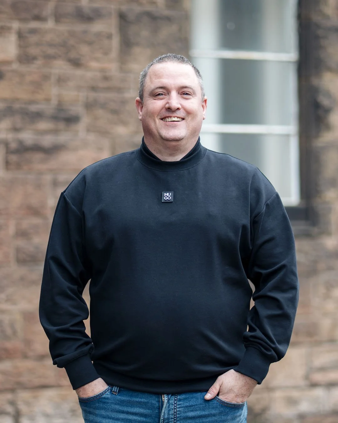 A smiling man with short hair wearing a black Hugo Boss sweatshirt and jeans, standing outdoors in front of a brick building with a window.