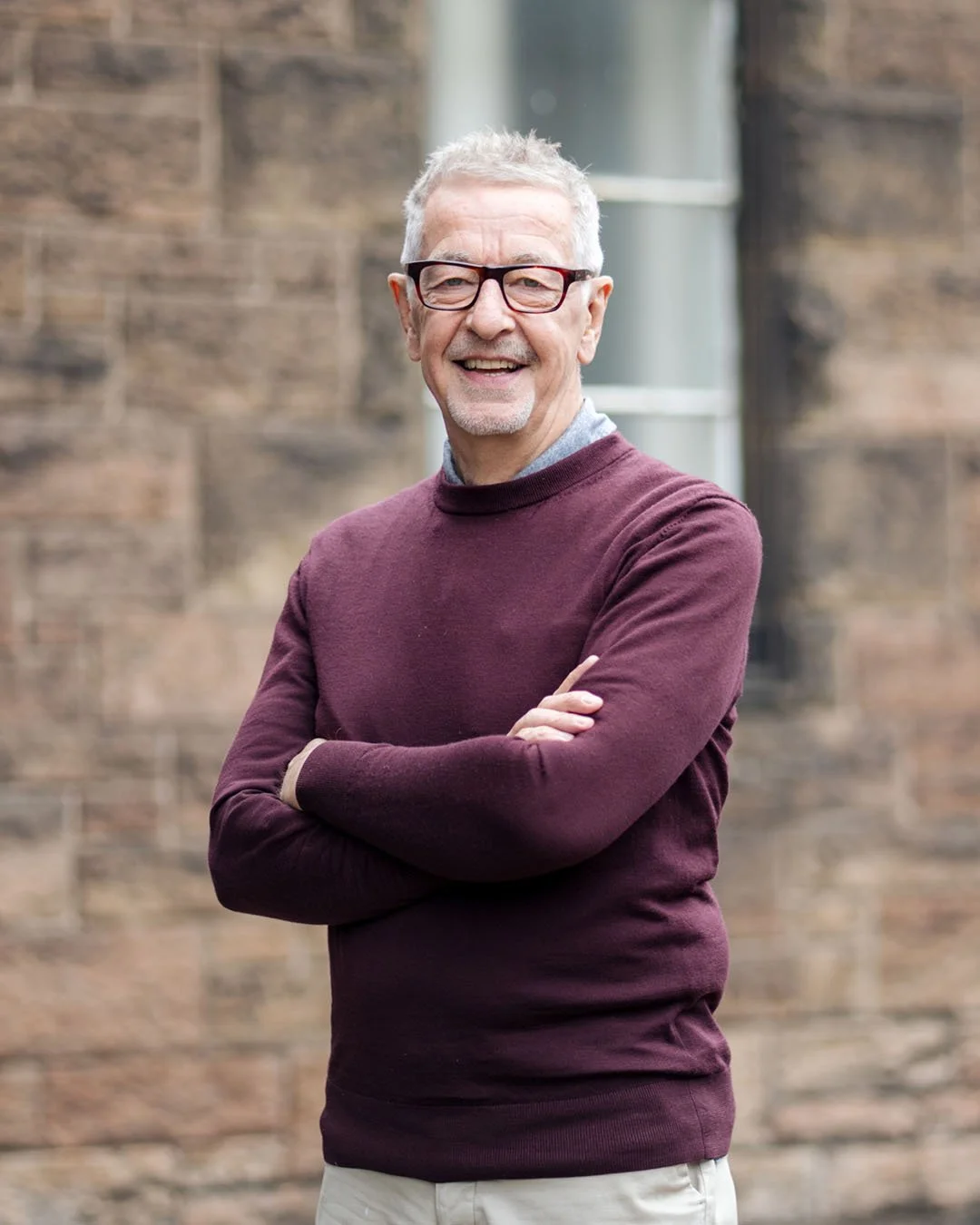 A cheerful older man with gray hair, glasses, wearing a maroon sweater over a gray shirt, standing outdoors with arms crossed in front of a brick wall.