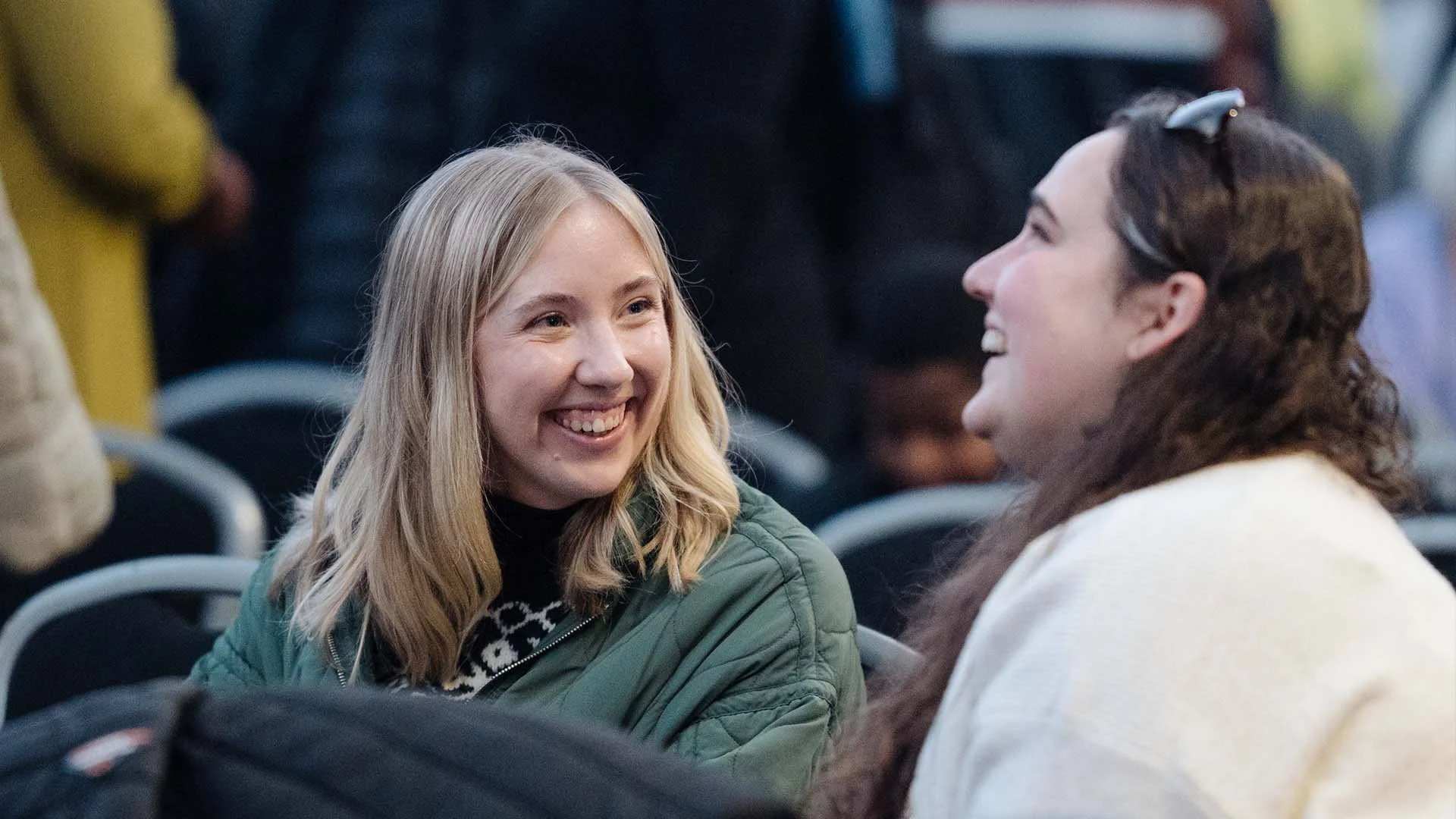 Two women smiling and talking at an indoor event, seated among other attendees.