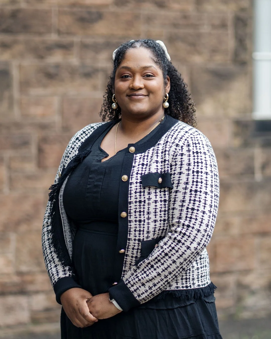 A woman standing outdoors in front of a brick wall, smiling, wearing a black dress and a navy and white patterned jacket with gold buttons and black trim, accessorized with earrings and a necklace.