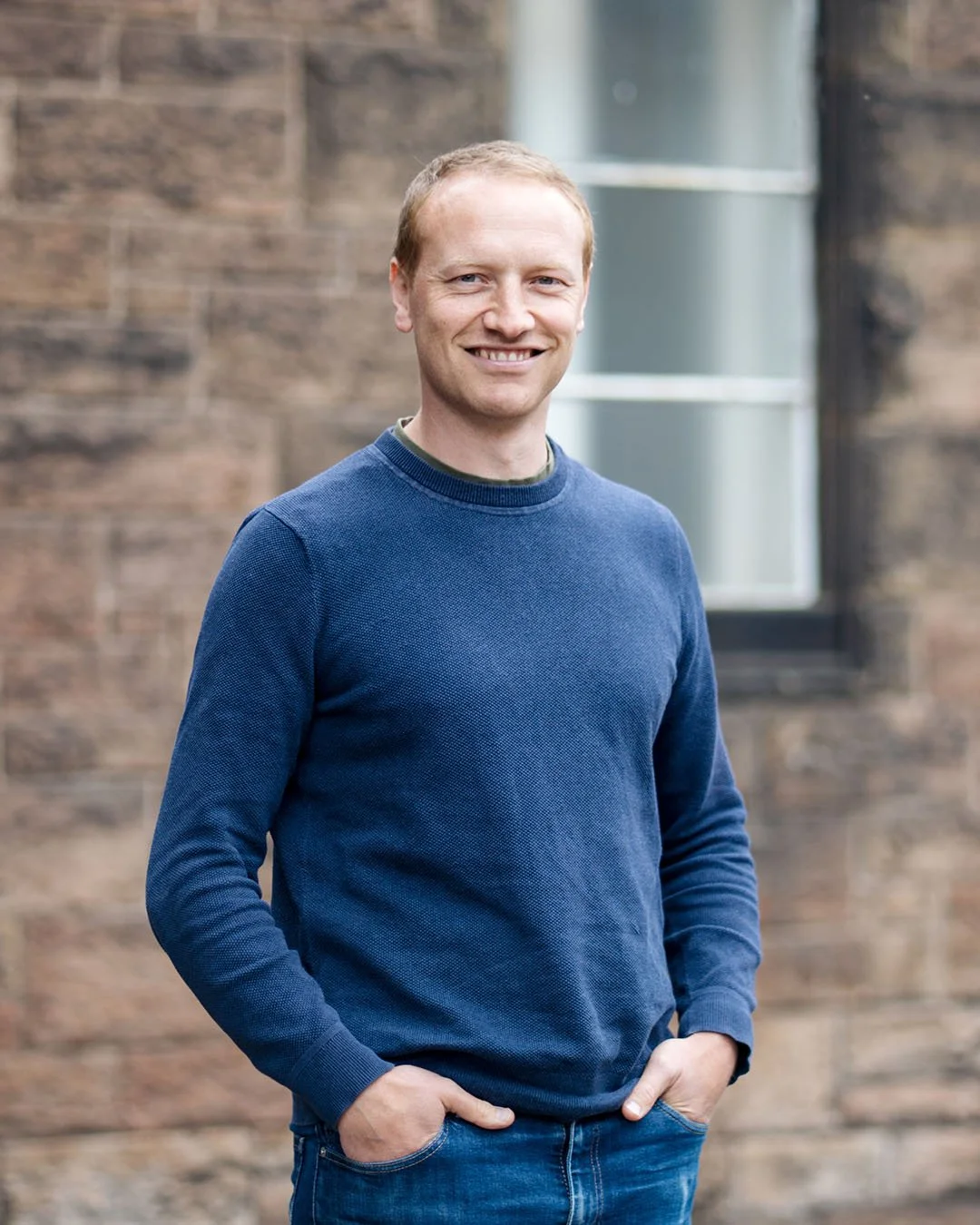 A man with short light brown hair and a light complexion smiling outdoors, wearing a navy blue sweater and jeans, standing in front of a brick building with a window.
