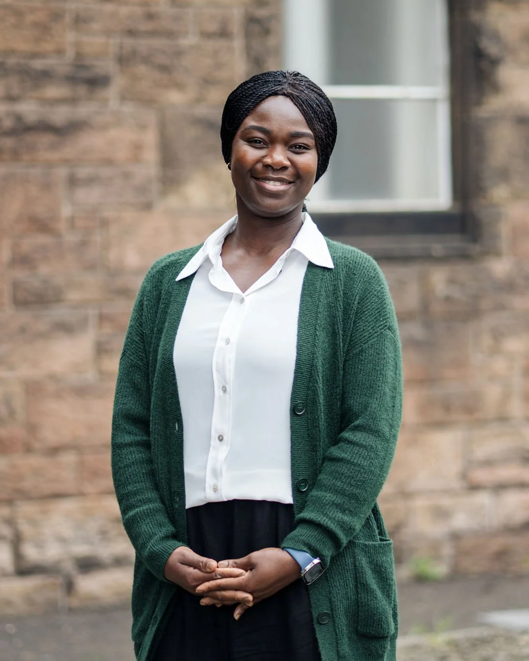A woman with braided hair smiling, wearing a white shirt and green cardigan, standing outside in front of a brick wall with a window.