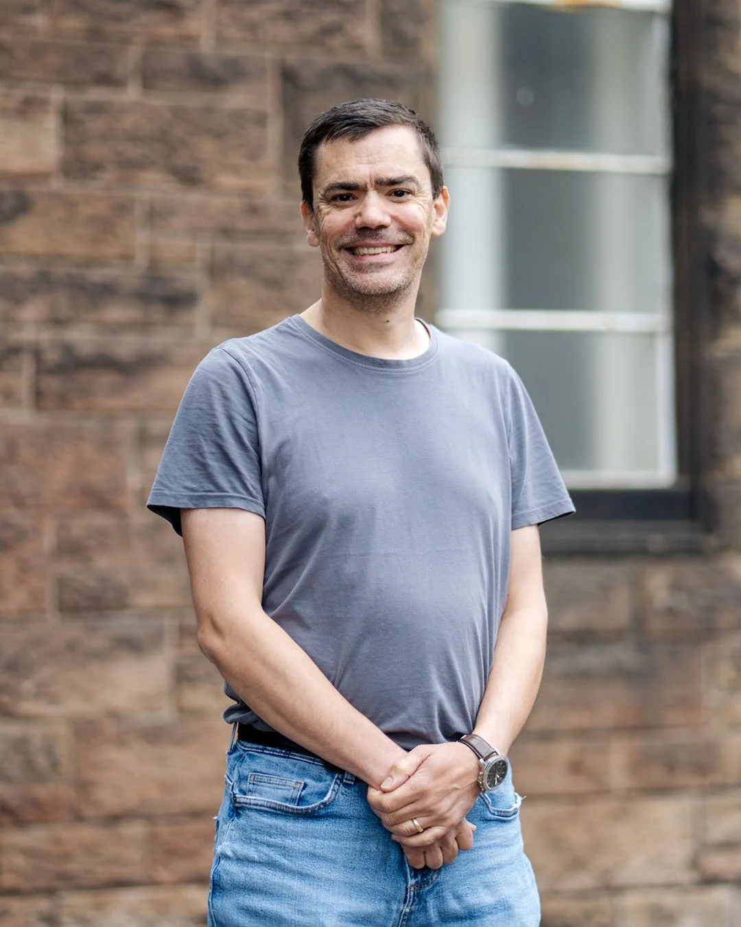 A man smiling outdoors in front of a brick building with a window, wearing a gray T-shirt and blue jeans.
