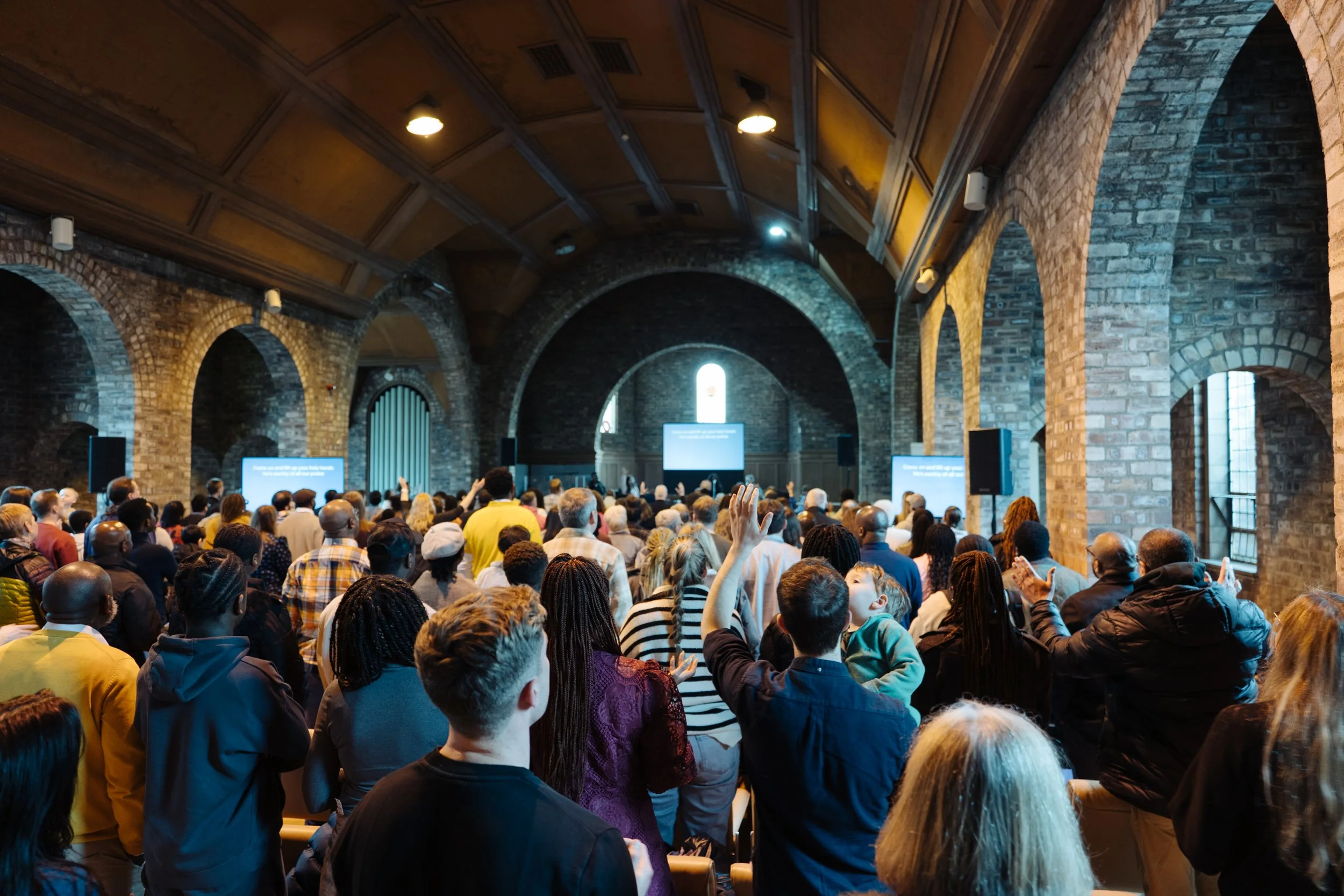 A large congregation of people gathered inside a historic brick church with a high arched wooden ceiling, facing a stage with a large digital screen.