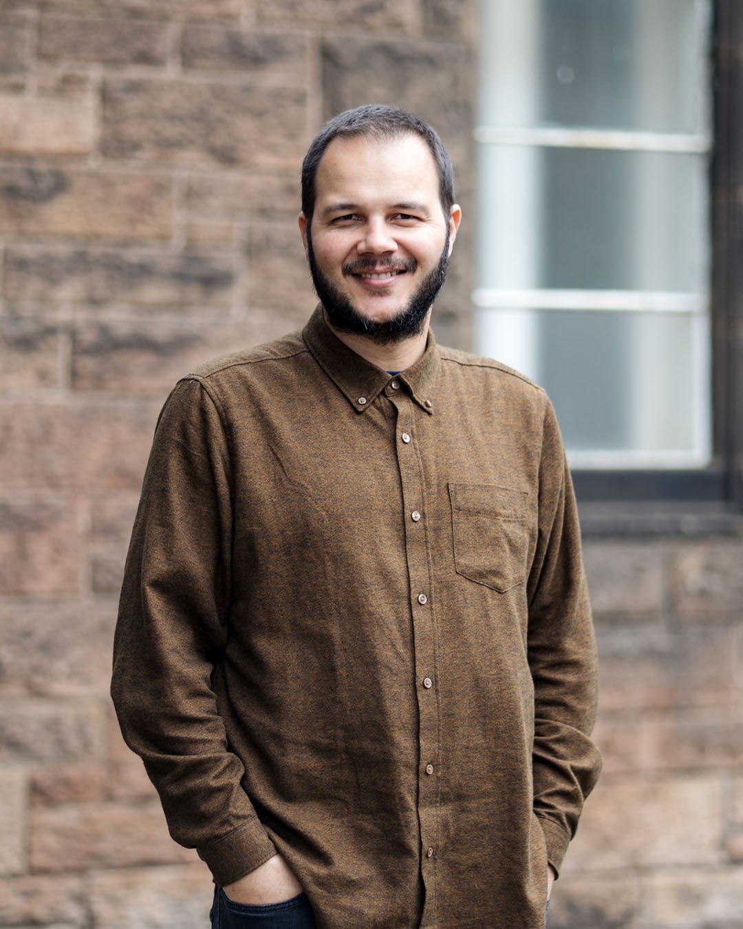 A man with a beard, wearing a brown button-up shirt, smiling outdoors with a brick wall and a window in the background.