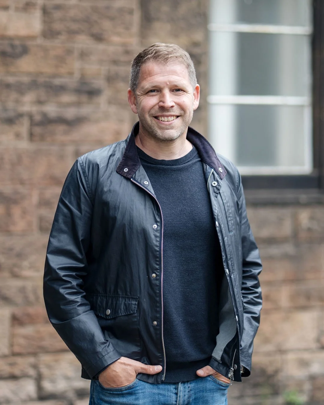 A smiling man standing outdoors in front of a brick wall and window. He is wearing a black leather jacket and blue jeans.