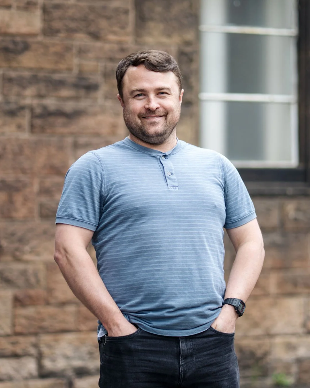 A man with short brown hair and a beard standing outdoors with hands in pockets, smiling, wearing a blue striped t-shirt and black jeans, in front of a brick wall and a window.