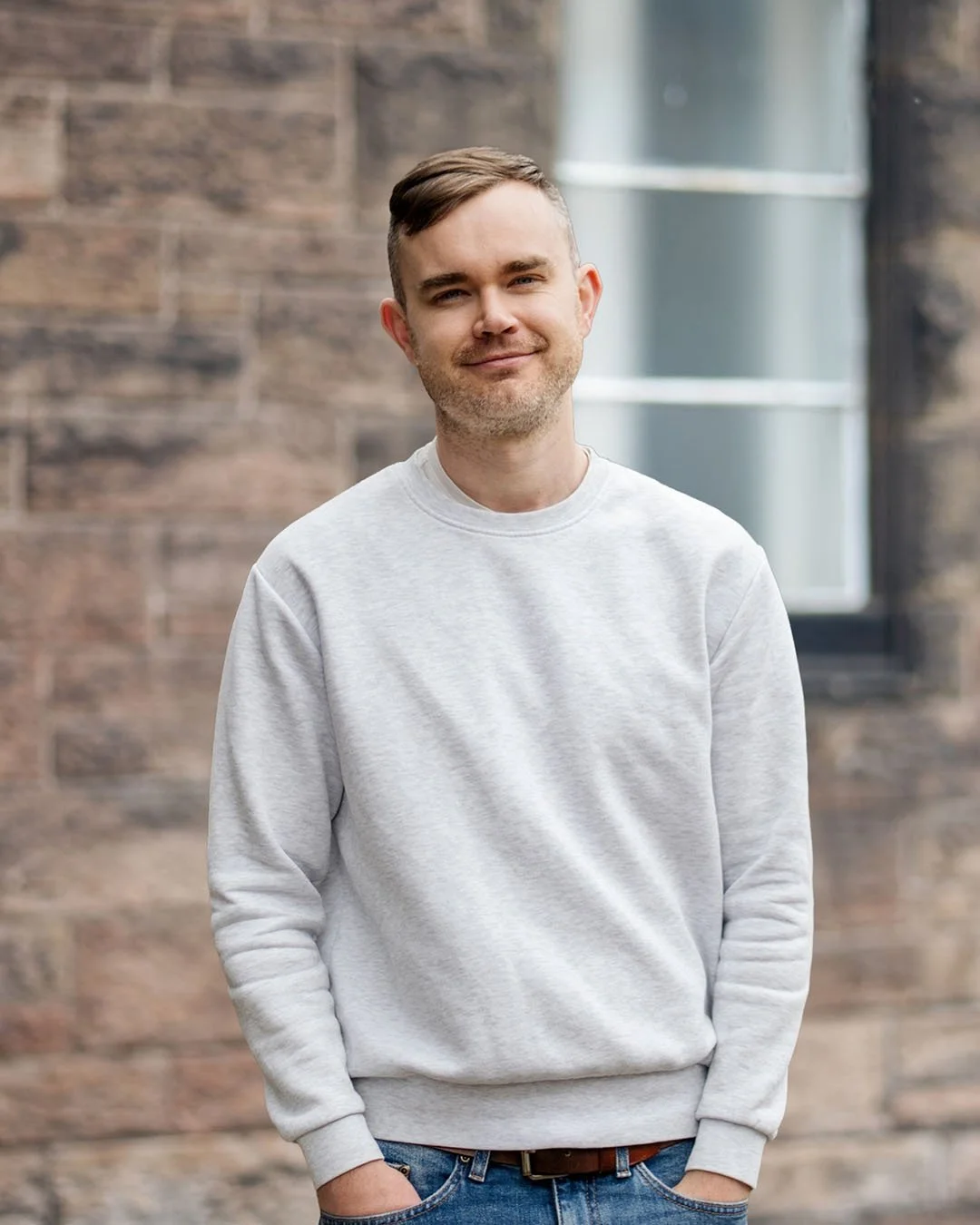 A young man with short brown hair and a light beard, wearing a light gray sweatshirt and blue jeans, standing outdoors in front of a brick wall and a window, smiling with hands in pockets.