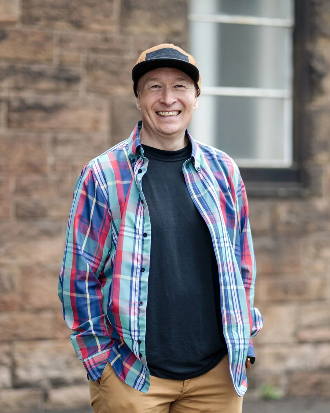 A smiling man wearing a checkered shirt, black T-shirt, tan pants, and a baseball cap, standing outdoors in front of a brick building with a window.