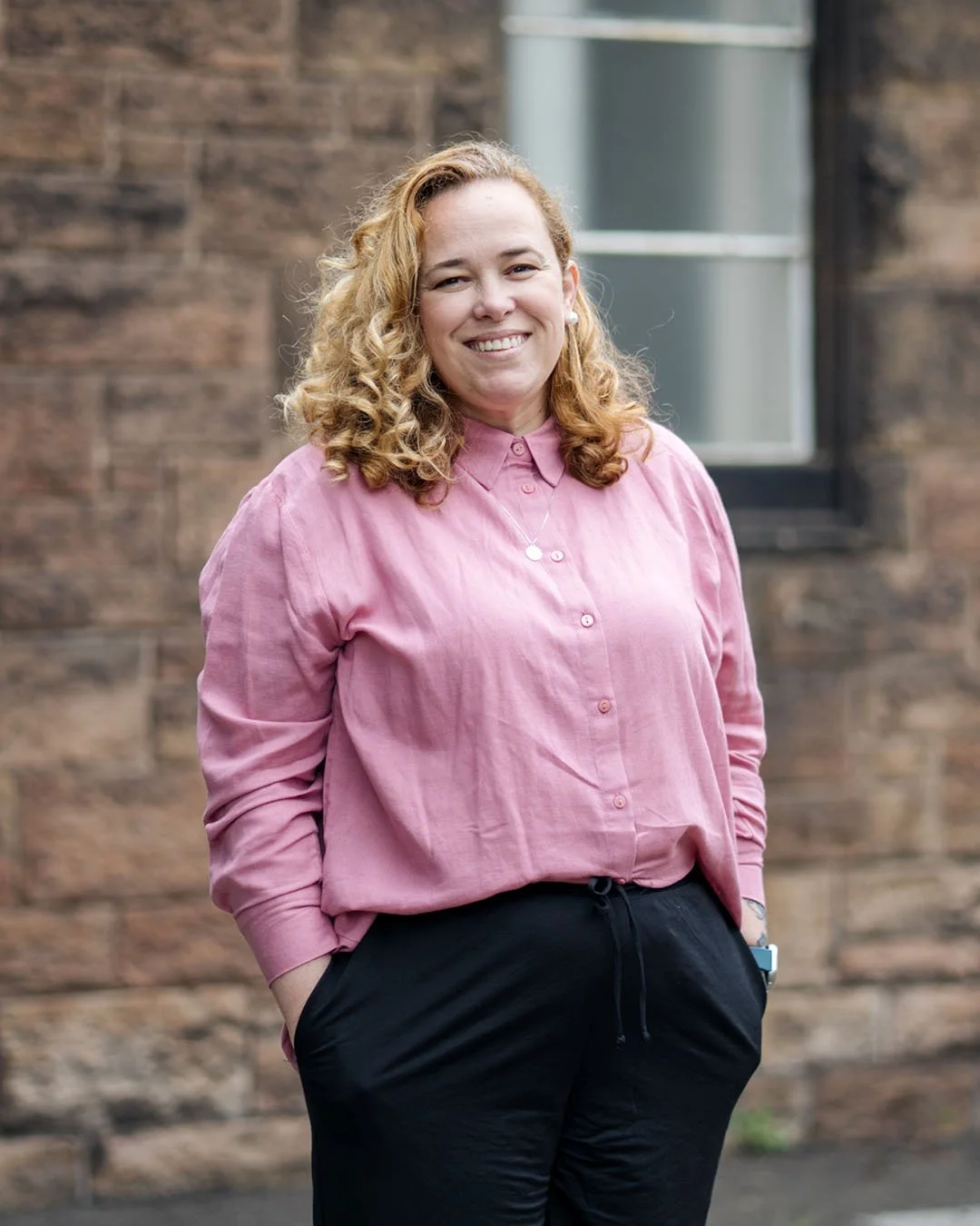 A smiling woman with curly blonde hair wearing a pink button-up shirt and black pants, standing outdoors in front of a brick wall with a window.