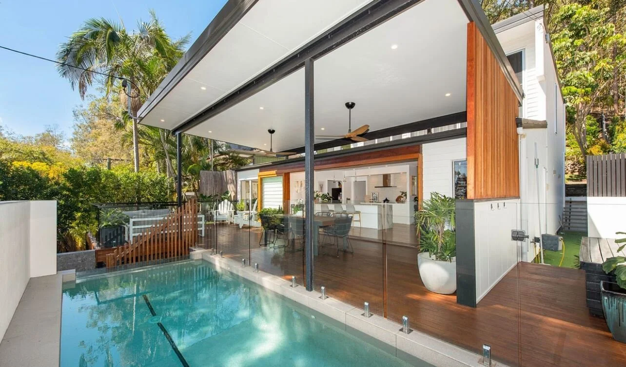 Modern backyard with a swimming pool, outdoor dining area, and a covered patio attached to a house with white siding and wooden accents, surrounded by lush greenery and palm trees.