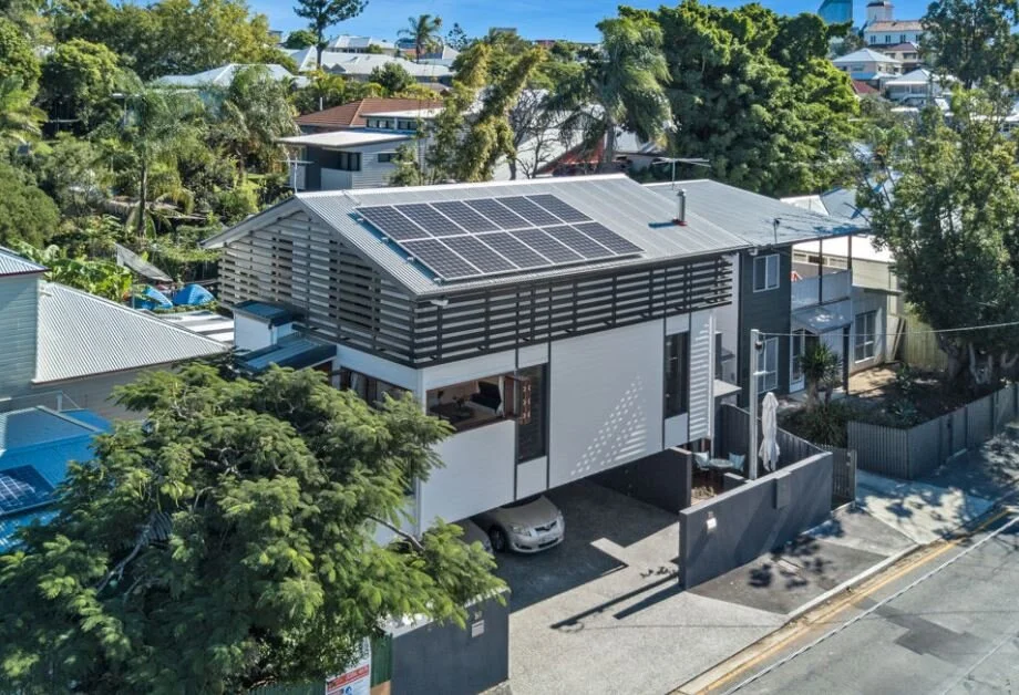 Modern two-story house with solar panels on the roof, surrounded by green trees in a residential neighborhood.