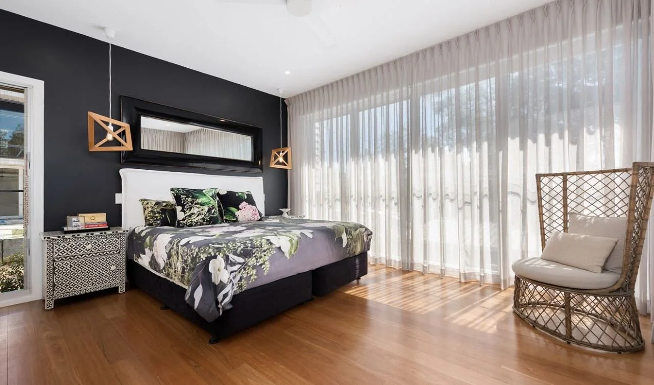A bedroom with a black accent wall, a white bed with floral bedding and pillows, a small patterned nightstand, a large mirror above the bed, floating wooden light fixtures, a chair with cushions, and sheer white curtains covering large windows.