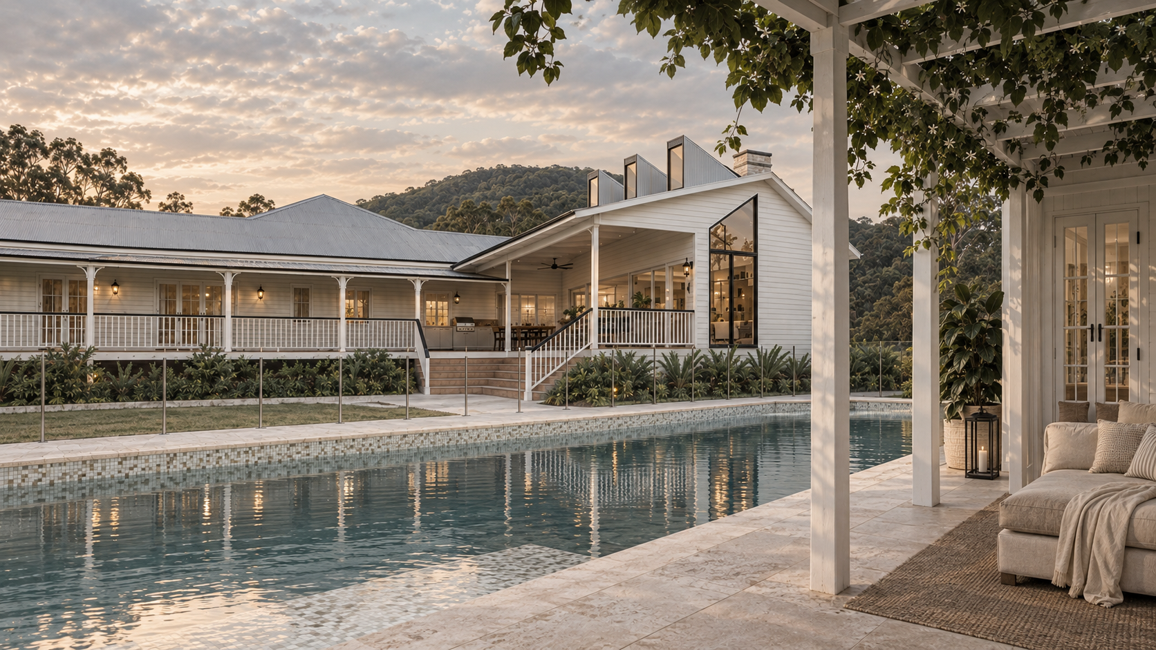 A luxurious house with a pool, outdoor seating area with a sofa and pillows, surrounded by greenery and hills, during sunset.